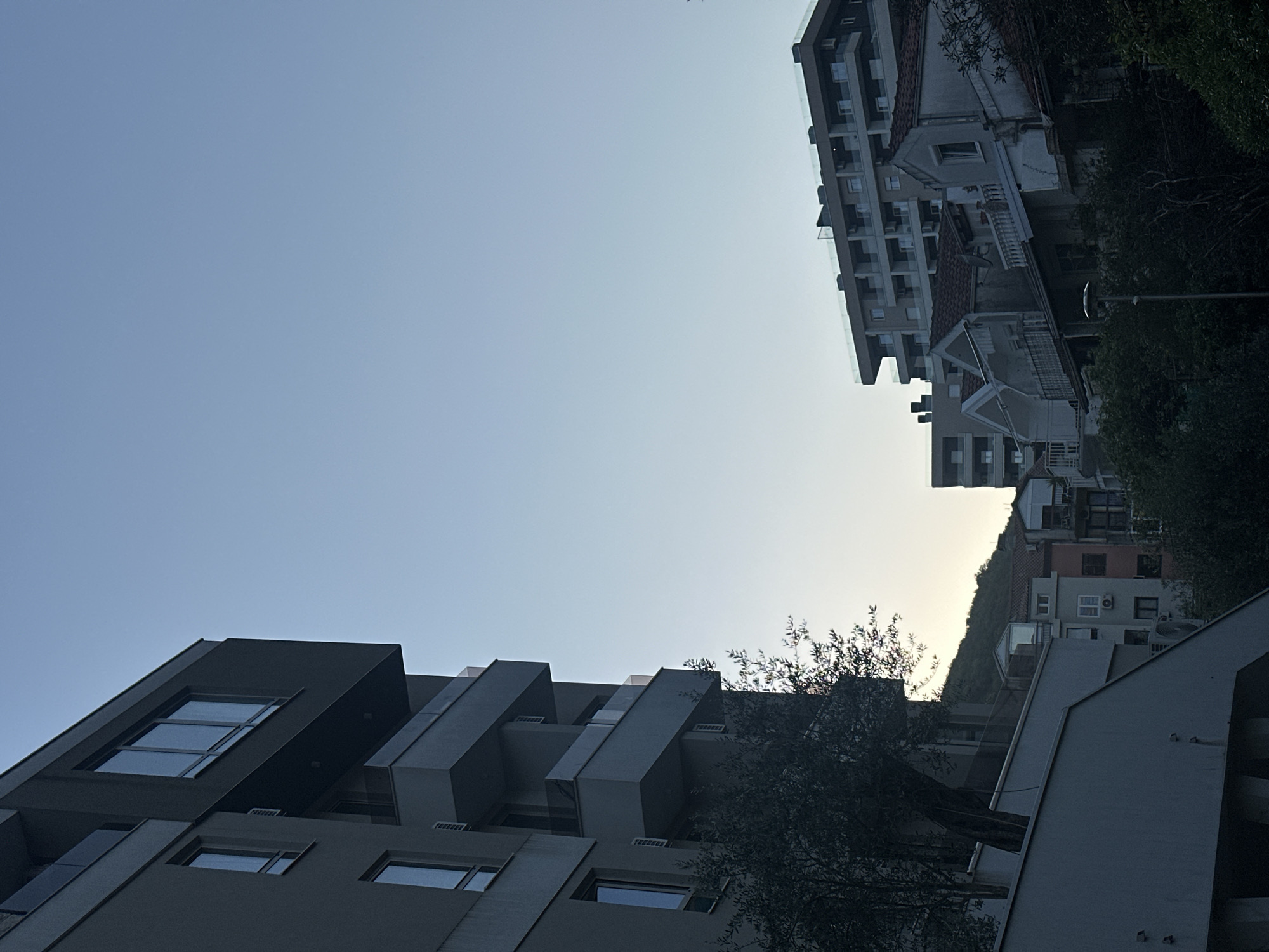 The image, when viewed in its correct orientation (rotated 90 degrees clockwise), captures an urban landscape in Budva, Montenegro, likely during sunrise or sunset. The sky, occupying the upper left quadrant, is a clear, light blue that gradually transitions to a pale, warm yellow-white hue towards the upper right, indicating the low position of the sun.
In the foreground on the left, a modern building with a dark grey or black facade stands prominently. It features angular, geometric designs with deep-set windows and cantilevered balcony-like structures, showcasing a contemporary architectural style. On the foreground right, a section of a tree with sparse foliage is visible, its branches reaching towards the sky.
To the right of the frame, another multi-story modern building with a lighter, possibly white or light grey, facade is visible. It features numerous balconies, many equipped with clear glass railings, suggesting an apartment complex or hotel.
Between and behind these modern structures, especially in the mid-ground and background on the right, several older, more traditional buildings are nestled. These feature classic terracotta-tiled roofs and lighter-colored facades, often with visible balconies and window details. They appear to be built into or on a lush green hillside densely covered with trees and vegetation, which ascends towards the horizon.
No people or discernible activities are taking place. The scene is static, emphasizing the architectural contrasts and natural light. The overall impression is one of a developing coastal town where modern construction coexists with older residences and natural landscapes. There is no visible text in the image.