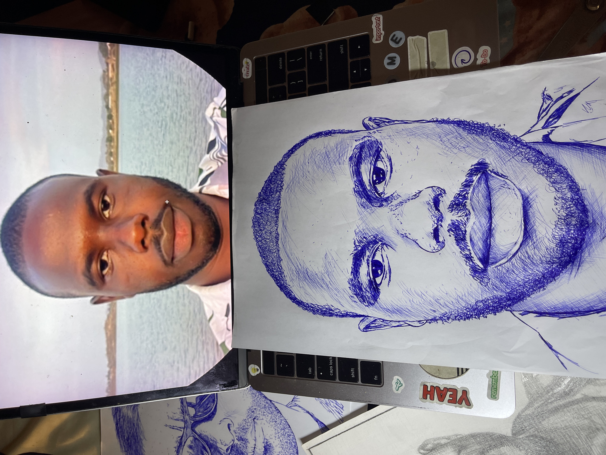 The media file displays a top-down view of a laptop with a drawing laid out on its keyboard and trackpad area. On the laptop screen, a portrait of a Black man is visible, positioned against a backdrop of water and distant foliage. To the right of the screen, a blue ink drawing of a Black man's face, rendered in a sketch style, is placed over a portion of the laptop. The drawing is detailed, with cross-hatching used to create shading and texture.
The laptop itself appears to be a MacBook, indicated by the keyboard layout and stickers on its surface. Visible text on the stickers includes "create," "important," "ME," "To-Do," "YEAH," and "remember." The drawing is on a white sheet of paper. The overall setting appears to be an indoor workspace. The lighting suggests it could be daytime, but there are no strong indicators of time of day or weather. The image captures a creative process, with a reference image on screen and a drawing in progress. The location context of Jos, Nigeria, suggests that the individuals depicted and the setting are likely associated with that region.