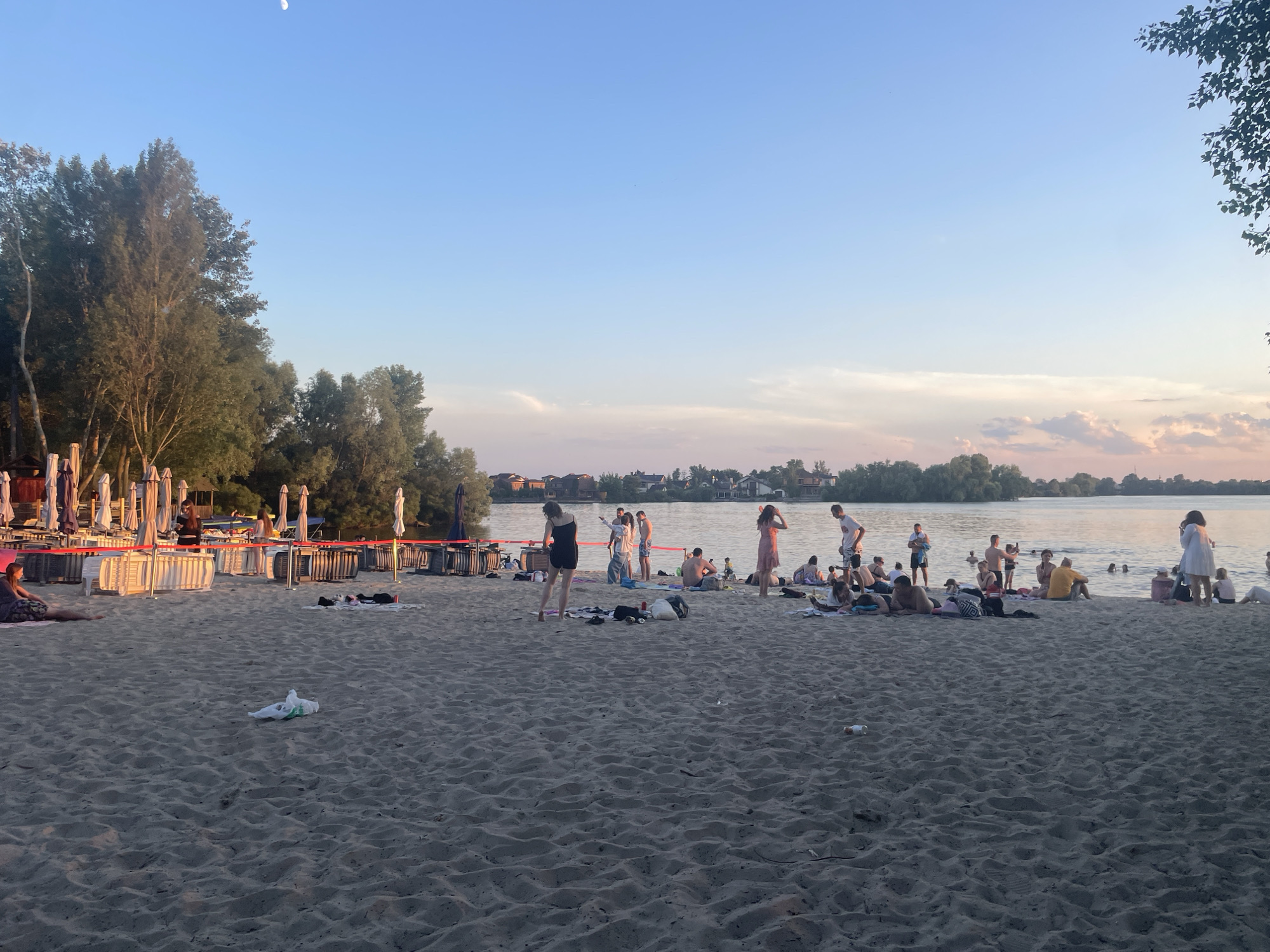 This is a daytime, outdoor, wide shot of a crowded sandy beach along a calm body of water, likely a river or lake, in Kyiv, Ukraine. The sky is clear and blue, with a faint crescent moon visible in the upper left corner. The sun is low, casting long shadows and bathing the scene in a warm, golden light, suggesting it is late afternoon or early evening.
A diverse group of people are engaged in various beach activities. Some are wading or swimming in the water, while others are relaxing on the sand, lying on towels, or sitting. A few individuals are standing and conversing. There are beach umbrellas and lounge chairs set up along the back of the beach, indicating a developed recreational area. Some litter, including a plastic bag and bottles, is visible on the sand. In the background, across the water, there is a shoreline with trees and buildings, suggesting a city or suburban area. The overall atmosphere appears relaxed and leisurely, with people enjoying the warm weather and the water.