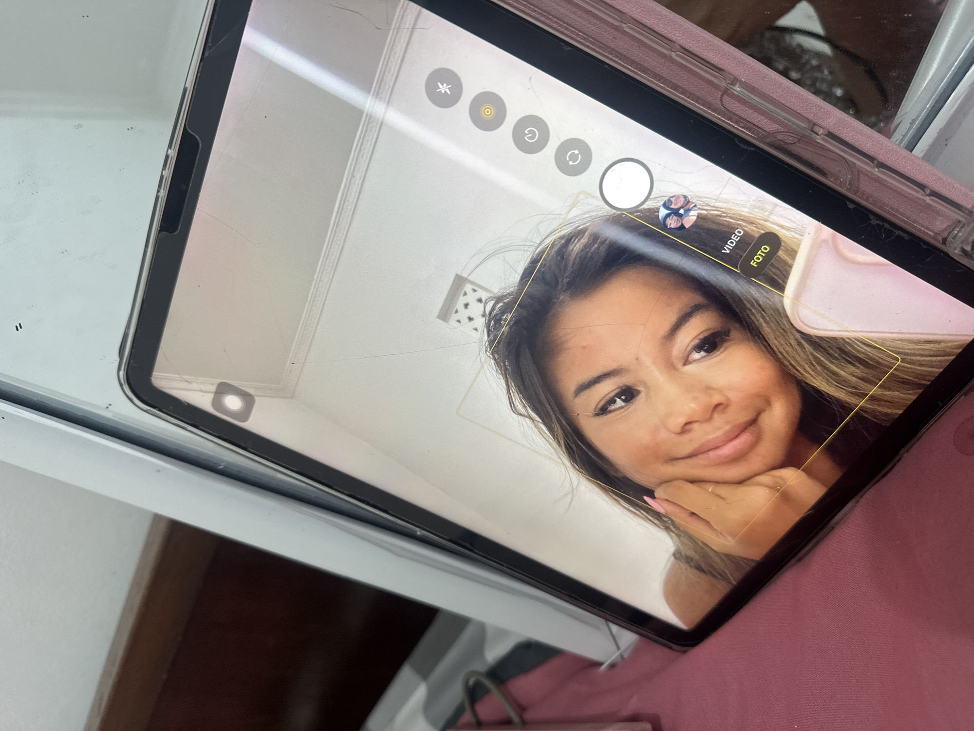 A person is visible on the screen of a tablet. The tablet is open to the camera application, which is actively displaying a selfie view of the person. The selfie shows a young adult female with brown hair and fair skin. She is smiling slightly with her chin resting on her hand. The background of the tablet screen is a white wall with a small ventilation grate. The tablet itself appears to be an iPad with a dark-colored bezel and a pink case. The tablet is positioned slightly askew, suggesting that the photo was taken hastily. The tablet is resting on a pink surface, presumably bedding or furniture. The image does not provide sufficient information to confirm the location in Sukawati, Indonesia.