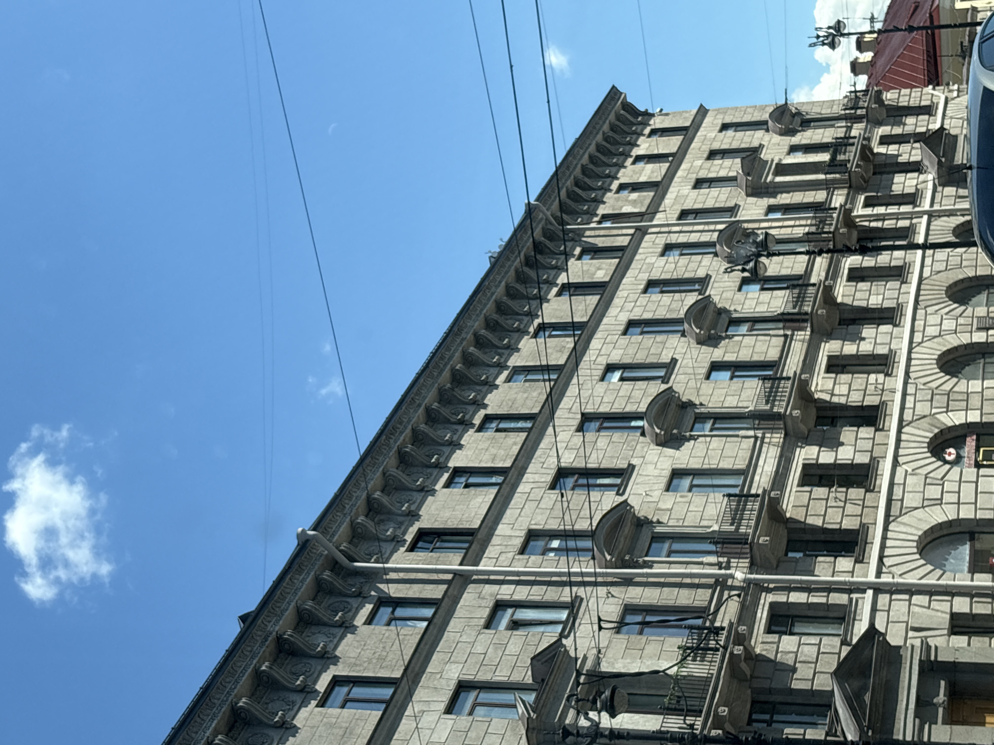 An exterior, eye-level, medium shot shows the facade of a multi-story neoclassical building on a clear, sunny day in Saint Petersburg, Russia. The building is constructed of light-colored stone blocks and features ornate decorative elements, including a heavily ornamented cornice and arched ground-floor entrances. Several windows, some with balconies, are visible across the facade. Overhead, utility wires crisscross the bright blue sky, punctuated by a few scattered white clouds. A street lamp is visible on the left side of the image, and the roof of another building with a red roof is partially visible on the right. The image captures the architectural grandeur typical of Saint Petersburg's historic city center.