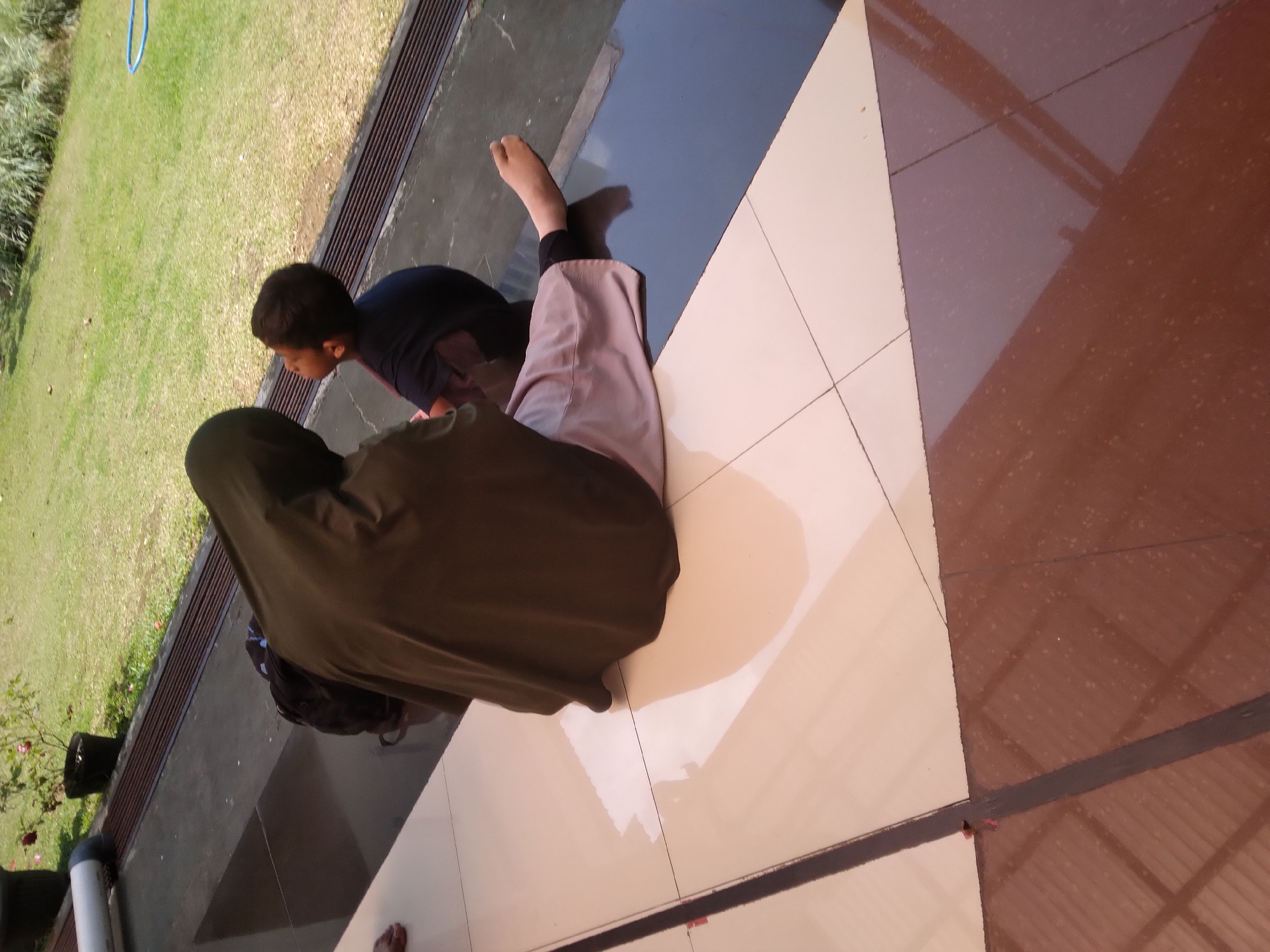 A person wearing a dark olive hijab and light purple pants sits on a tiled floor, facing away from the camera, with their legs extended. To their right, a young boy in a dark blue shirt looks upwards. They are positioned on a tiled patio area adjacent to a grassy lawn. A dark backpack is visible behind the person in the hijab. In the background, a white pipe and a potted plant with red roses are present, along with a dark, slatted drainage channel. The environment suggests an outdoor setting in what is likely a residential or public space. The image is taken during daylight hours, with light reflecting off the polished tiles. The location is identified as Desa Pamijen, Kecamatan Sokaraja, Indonesia.