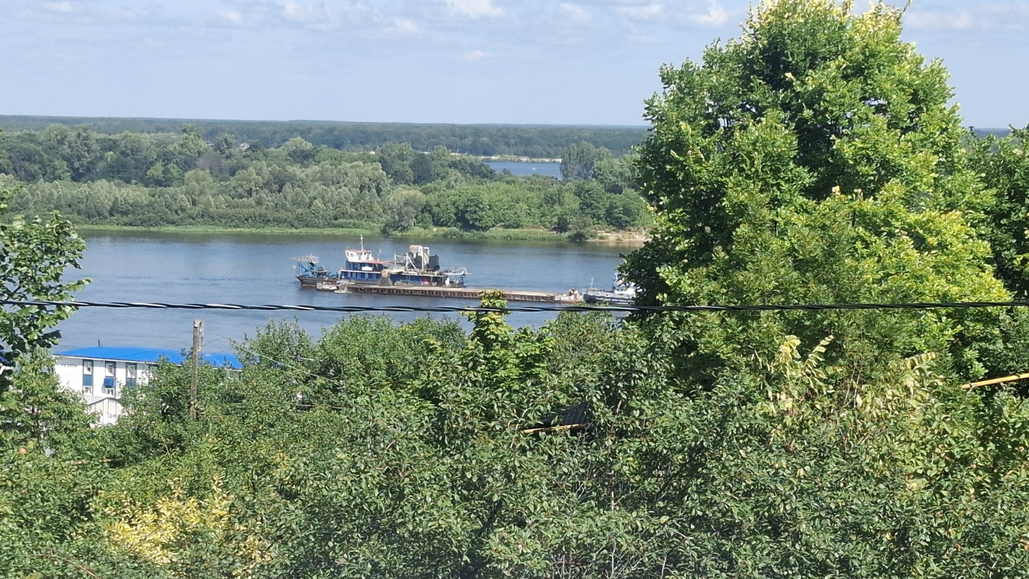An eye-level, medium outdoor shot captures a wide, calm river under a partly cloudy blue sky. A multi-part barge, including what appears to be a dredging or construction vessel, is positioned in the middle of the river. The riverbank opposite the viewer is densely wooded with a mix of deciduous trees. Closer to the viewer, on the left side of the frame, a building with a bright blue roof and white siding is partially visible, situated amongst lush green foliage. A prominent, mature tree with vibrant green leaves dominates the right side of the image, its branches extending towards the center. Black wires are visible crisscrossing the foreground, obscuring some of the lower foliage and the building.