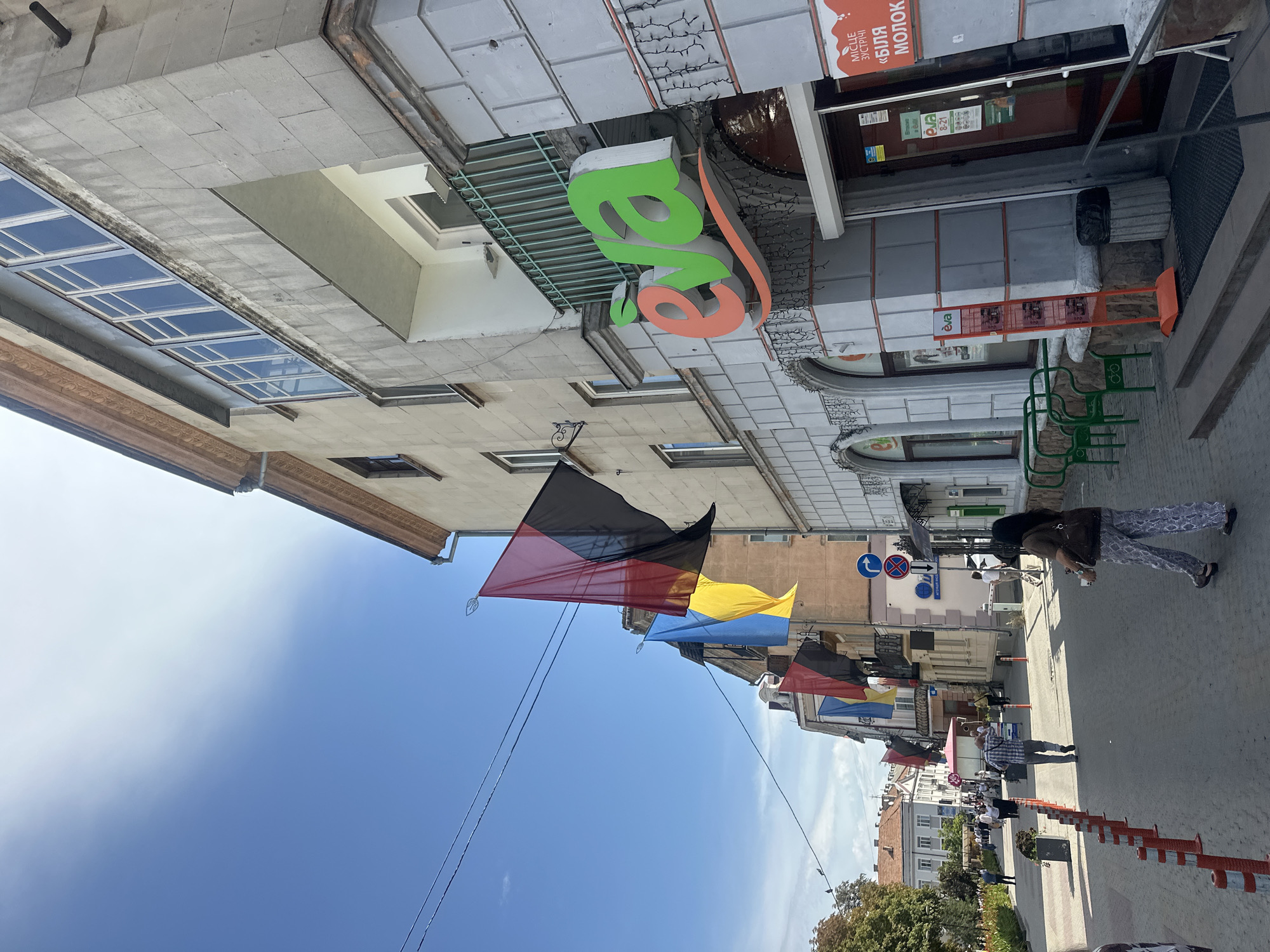 The image shows a street in Ternopil, Ukraine, with several flags displayed on buildings. The flags appear to be black and red, possibly representing Ukrainian national or historical symbolism, alongside a blue and yellow Ukrainian flag. People are walking on the sidewalk, and there are shops and businesses visible. One shop has a prominent sign with the name "eva" in colorful lettering. The architecture suggests an urban environment with multi-story buildings. The sky is clear and blue, indicating a sunny day. There are orange traffic cones and bollards lining the sidewalk, and some greenery in planters.