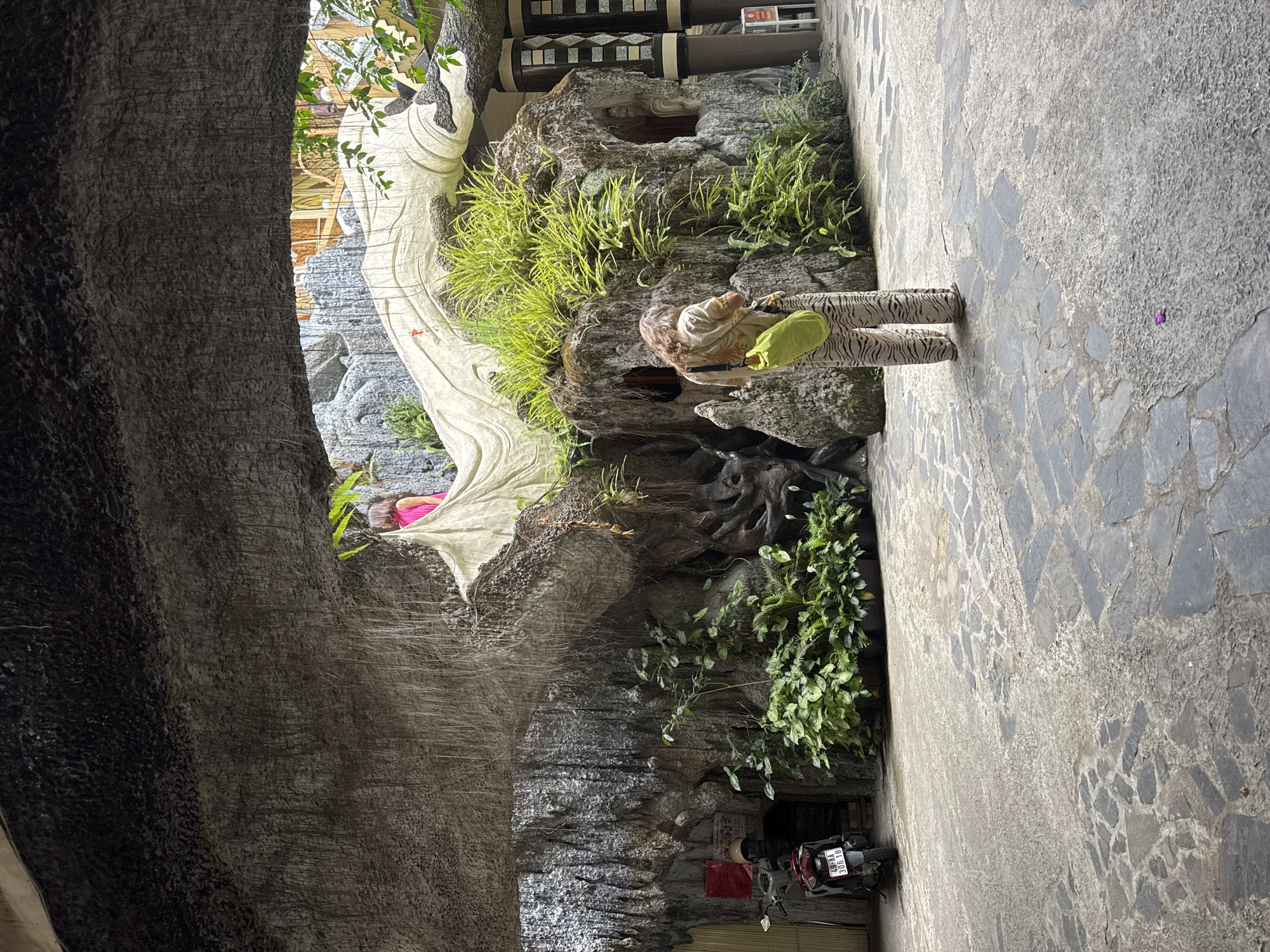The image depicts the exterior of the Crazy House (Hang Nga Guesthouse) in Da Lat, Vietnam. In the foreground, a person wearing tiger-striped pants and a light-colored shirt with a green bag slung over their shoulder stands facing a large, organically shaped rock structure covered in lush green vegetation. Behind this person, part of the iconic, winding architecture of the Crazy House is visible, including a sculpted, large white form resembling a tree trunk or flowing drapery. In the background, another person in a pink top is visible on an elevated walkway. To the left, a red and silver scooter is parked. The ground is paved with irregular flagstones. The overall scene suggests a unique and fantastical architectural space integrated with natural elements.