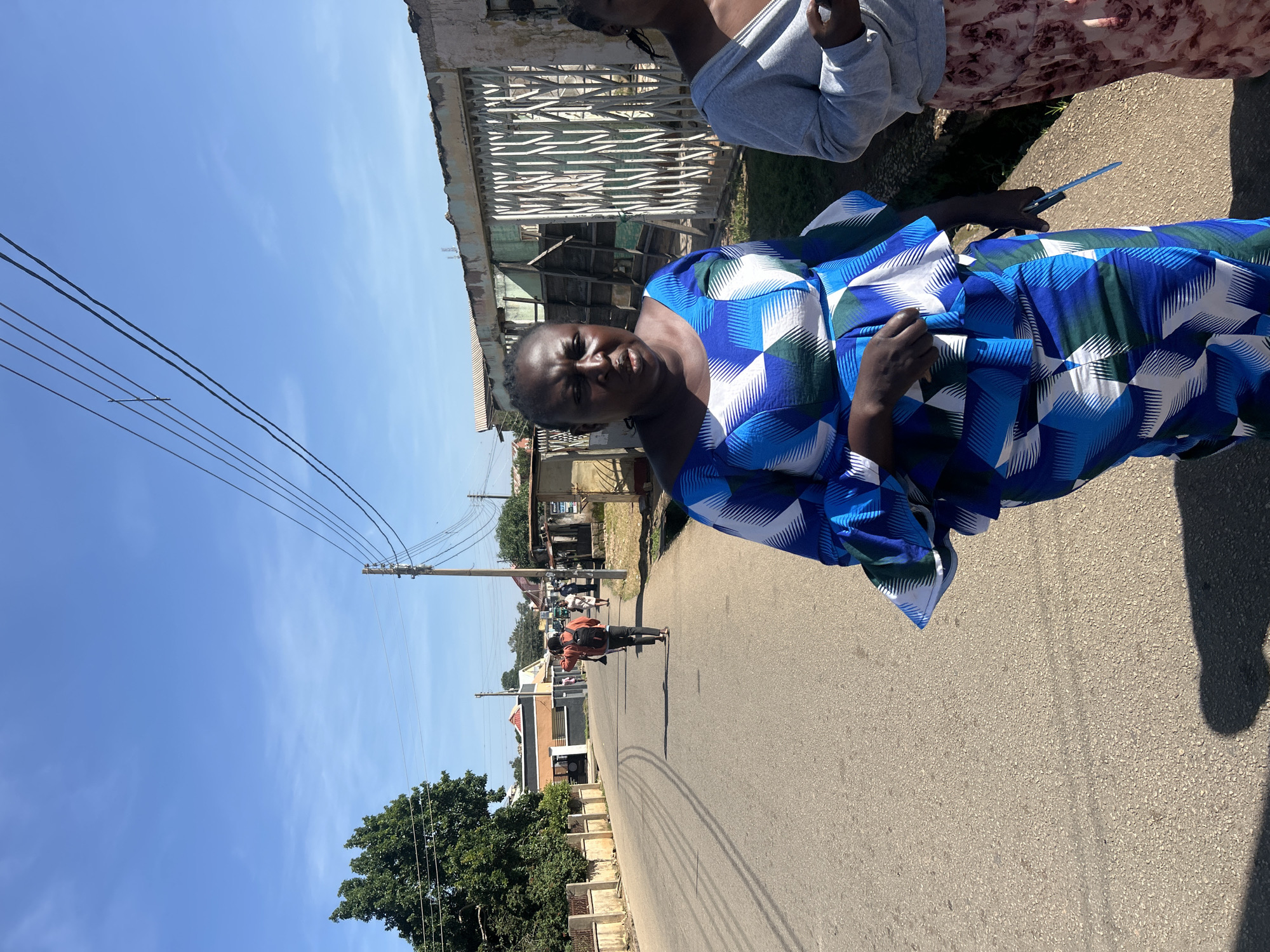The image shows a woman in the foreground wearing a blue and white patterned dress, walking on a street. She is looking towards the camera. Behind her, another woman in a grey hoodie and a patterned skirt is partially visible. In the background, a person with a backpack is walking away from the camera down the street. The street is paved and lined with buildings and trees. Power lines are visible overhead. The scene appears to be in a city, possibly Jos, Nigeria, given the architectural styles and vegetation. The overall impression is a bright, sunny day.