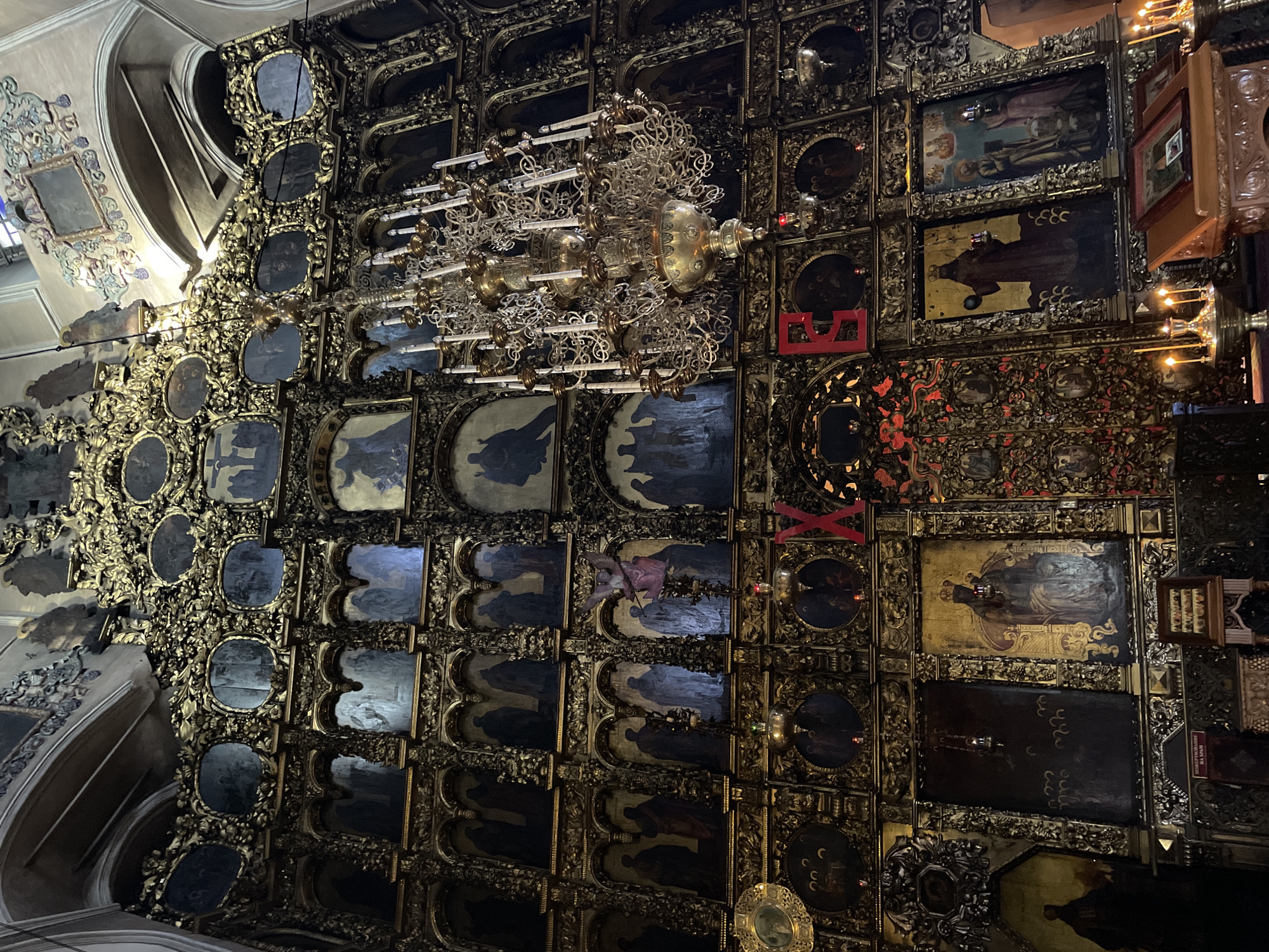 The image presents a detailed, upwards-looking view of an exquisitely ornate interior, likely the ceiling or a towering wall within a church, cathedral, or monastery. The surface is densely covered with numerous religious icons, predominantly rectangular and oval in shape, set against a backdrop of intricate golden carvings and decorative elements.
The icons themselves feature dark, muted backgrounds, with figures depicting saints or biblical scenes rendered in somber tones of dark blue, brown, and grey. These paintings are framed by an elaborate network of gilded filigree, scrolls, and floral patterns that fill almost every available space, creating a sense of overwhelming opulence and detail.
A large, multi-tiered chandelier, crafted from gold-colored metal with numerous ornate branches and light fixtures resembling candles, hangs prominently in the upper-middle section of the frame. Smaller, individual lamps, some with red backings, are strategically placed among the icons, casting warm light that highlights the gilded surfaces.
Towards the upper left corner, a lighter-colored architectural feature, possibly an arch or part of a dome, is visible, adorned with more subdued, pastel-colored decorations. The dominant aesthetic is a rich contrast between the dark, profound imagery of the icons and the radiant, intricate golden craftsmanship. Red accents are also subtly integrated into the decorative scheme. No people are visible, allowing the viewer to focus entirely on the architectural and artistic splendor. A small white sign with black Cyrillic text, partially visible in the bottom right, reads "ВАЖНА ИНФОРМАЦИЯ НА ХРАМ," translating to "IMPORTANT INFORMATION IN THE TEMPLE/CHURCH."