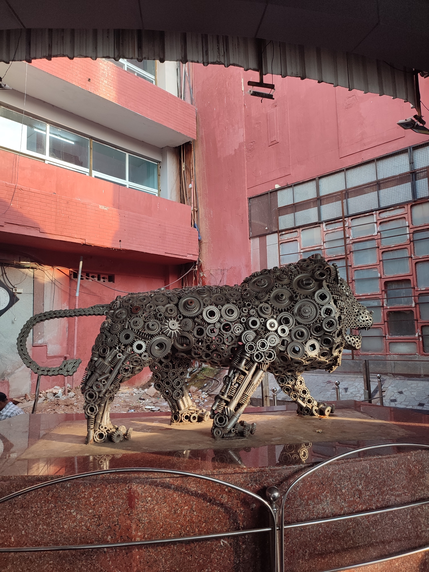 Here's a detailed description of the image:
The image shows a metal sculpture of a lion, constructed from gears, cogs, and other mechanical components. The sculpture is positioned on a red marble-like platform, surrounded by a metal railing with rounded posts.
The backdrop consists of a red brick building, with a section of windows. To the left, there are some power lines and a person. The upper part of the image is partially obscured by a corrugated structure.
The location is Unknown, Unknown.