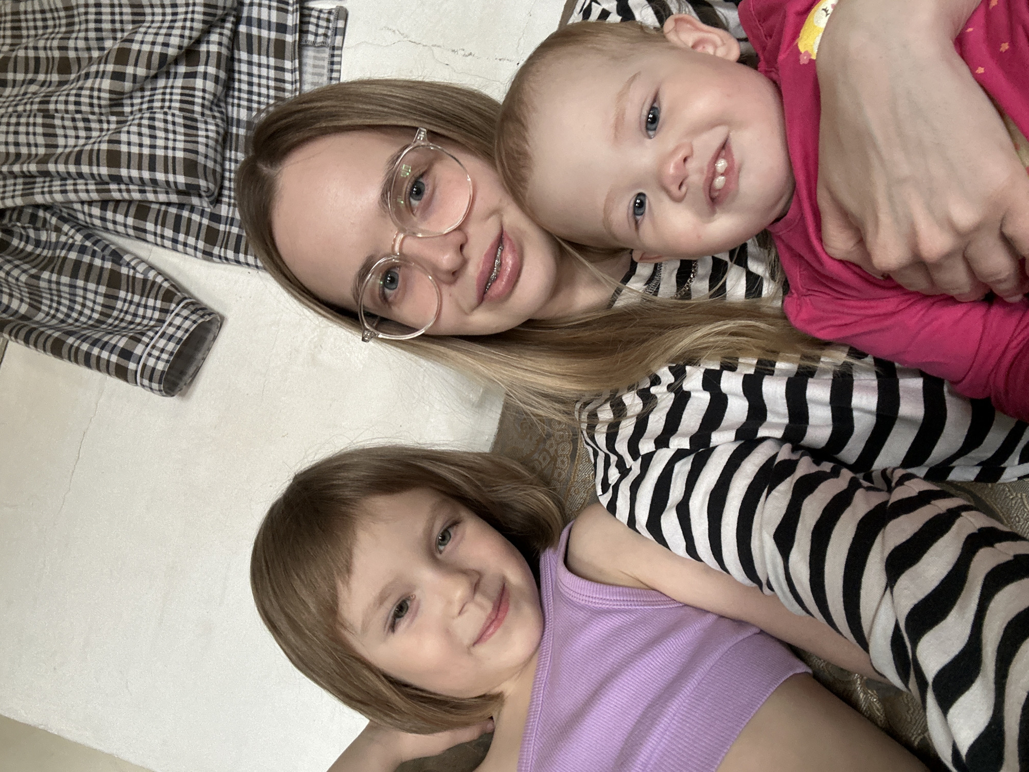The image shows a selfie-style view of a woman with two children. The woman is wearing glasses and braces, with long blonde hair. A toddler in a pink shirt is held close to her face, smiling. A young girl is lying below the woman, also smiling. The woman is wearing a striped shirt. Part of a plaid garment is visible in the upper left corner of the image. It is impossible to verify the location of the people in the image.