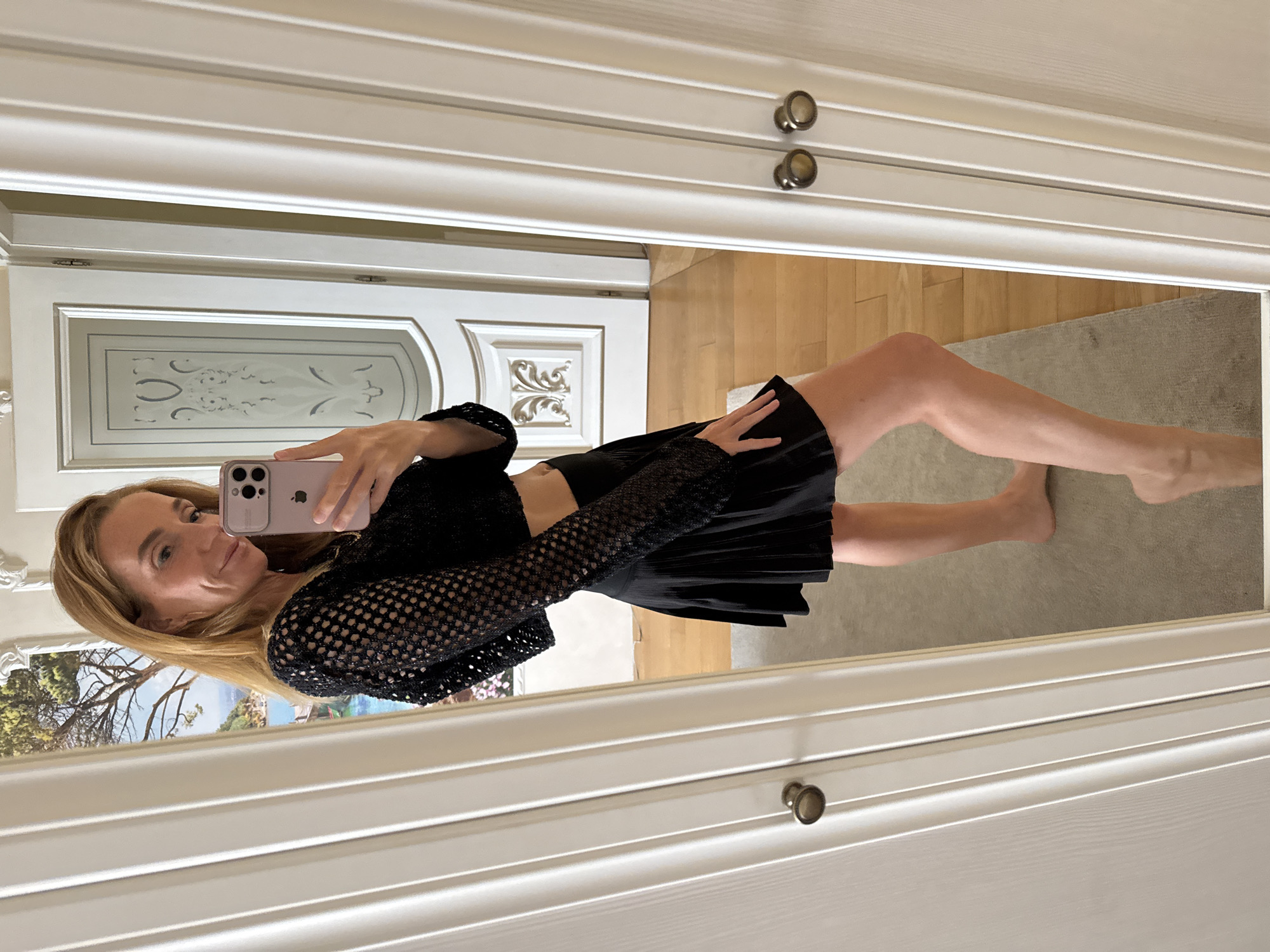 A woman is taking a selfie in a mirror, wearing a black, sparkly, mesh crop top and a black pleated skirt. She has blonde hair and is smiling. Her phone, a pink iPhone, is visible as she holds it up to take the photo. The background shows a mirrored wardrobe with white doors and ornate detailing. A painting of a coastal landscape with trees and water is reflected in the mirror behind her. The floor appears to be made of light-colored wood. The scene is indoors, likely in a bedroom or dressing area. The lighting is bright, suggesting it is daytime.