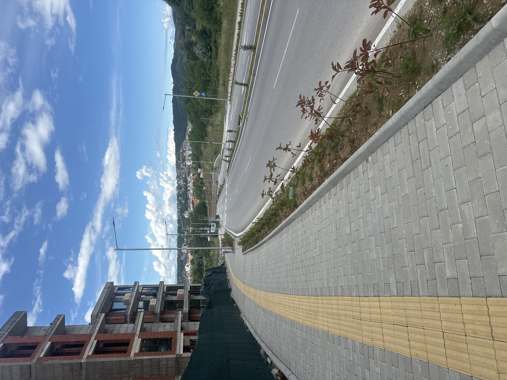 The image displays a daytime scene in Blagoevgrad, Bulgaria, featuring new urban infrastructure and an ongoing construction project under a bright blue sky with scattered white clouds.
In the left foreground, an unfinished multi-story building is visible, constructed with concrete frames and red brickwork, with some dark-paned windows already installed. A dark green construction net or tarp covers the lower section of this building.
Adjacent to the construction site and extending into the midground is a wide, newly paved sidewalk made of grey rectangular interlocking pavers. A prominent yellow tactile paving strip runs down the center of this sidewalk, designed to assist visually impaired pedestrians. A grey curb separates the sidewalk from a narrow, planted median strip containing small, red-leafed plants.
Beyond the sidewalk and median lies a modern, multi-lane asphalt road with distinct white lane markings. Another median strip, featuring small green saplings and street lamps, divides the road lanes. In the midground, a dense green hillside rises, behind which a sprawling town or city with numerous buildings can be seen, nestled in a valley. Further in the background, more green hills stretch towards the horizon.
The sky is clear and bright, indicating midday or early afternoon. Visible details include street lamps along the road and several road signs; one notable sign is a blue square with a yellow diamond-shaped pedestrian crossing symbol and another yellow diamond with a right-pointing arrow. No people are present in the scene.