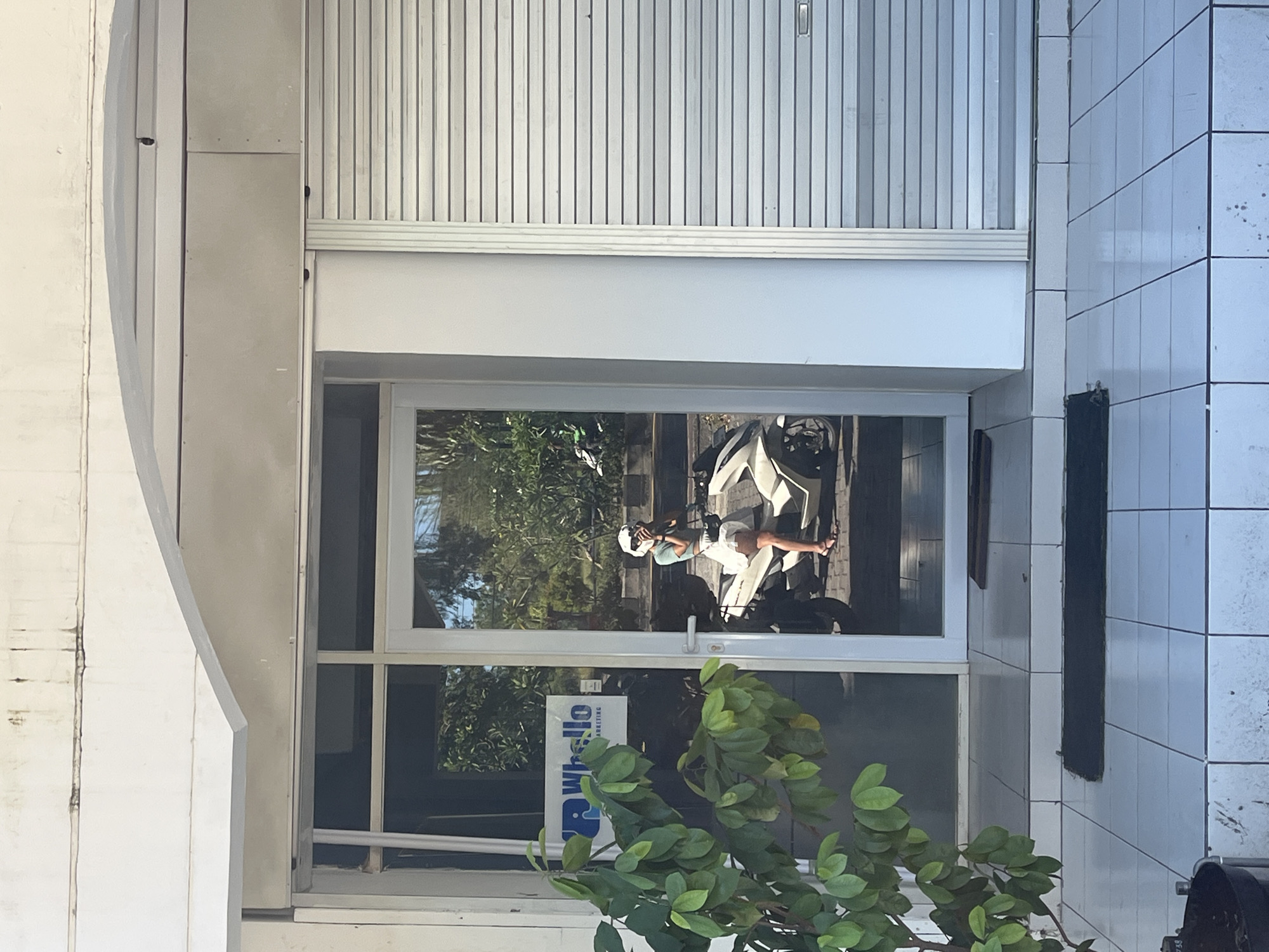 A horizontally oriented outdoor shot shows a building facade with a closed metal roller shutter at the top, a white frame window below it, and tiled walls on the right side.
The window reflects a sunlit exterior scene with trees and a person lounging on a white chair. The person is wearing a white helmet, a light-colored top, and shorts. A logo for "Whella Marketing" is visible on the window's lower portion, obscured by a leafy green plant in front.
The tiled wall to the right features rectangular white tiles and a dark vertical element fixed to the surface. The bottom right corner of the image includes a partial view of an object that appears to be a trash receptacle.
The image appears to have been taken during daylight hours, based on the light and shadows. The location is Kuta Selatan, Indonesia, implied by the local business name "Whella Marketing".