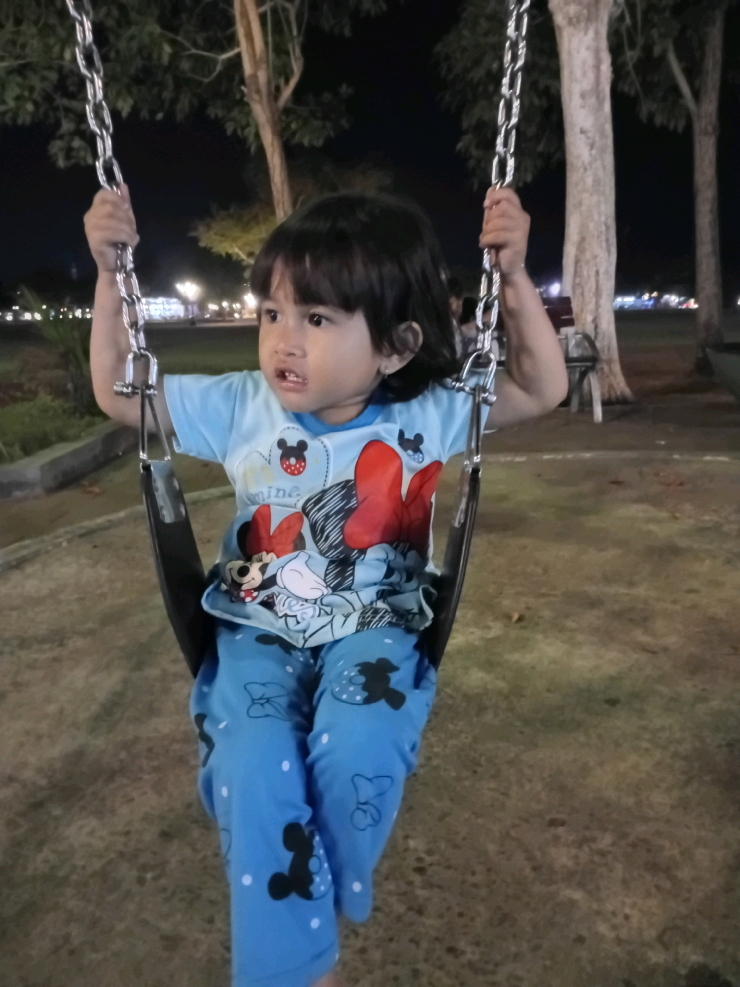 A young child is seated on a swing in an outdoor park setting at night. The child is wearing a light blue t-shirt with a Minnie Mouse graphic and blue pants with a Minnie Mouse pattern. The child's hands are gripping the chains of the swing. In the background, there are trees and distant lights, suggesting an urban or park environment. The ground appears to be paved or covered in dirt. The overall lighting suggests it is nighttime, with artificial light sources illuminating the area.