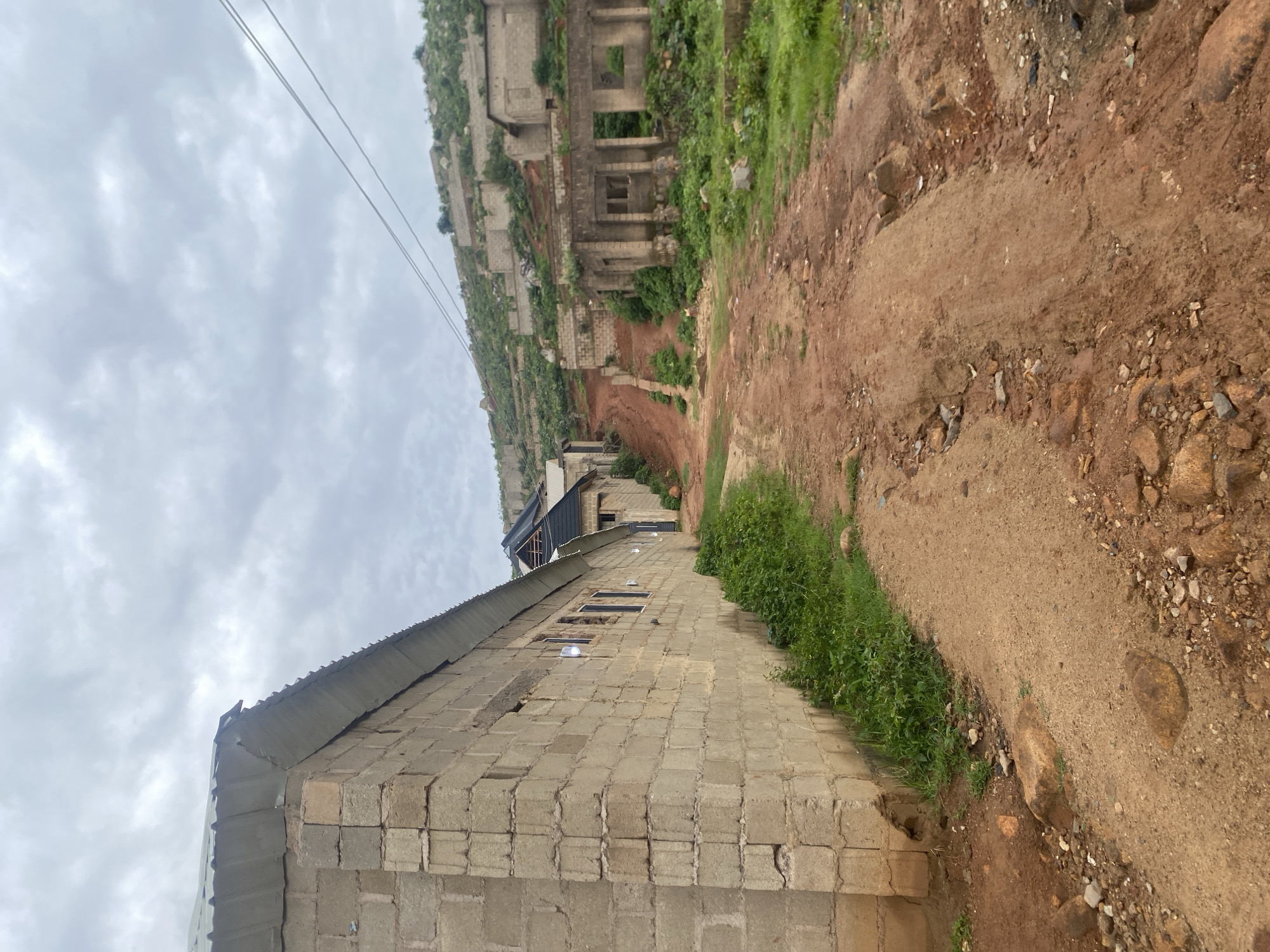 The image depicts a scene in a hillside urban area in Jos, Nigeria. In the foreground, a dusty red dirt road winds upwards. To the left, a long, unfinished building constructed of cinder blocks runs along the road. Its corrugated metal roof is visible, extending past the block walls. Small green plants grow along the base of this building. Further up the road, more structures are visible, including some that appear to be in a state of disrepair or are unfinished, with exposed walls and roofs. These structures are scattered across the rocky, vegetated hillside. The sky is overcast with heavy clouds. Power lines are visible in the upper right portion of the image, stretching across the sky above the hillside. The overall impression is one of an ongoing or abandoned construction site on a challenging terrain.