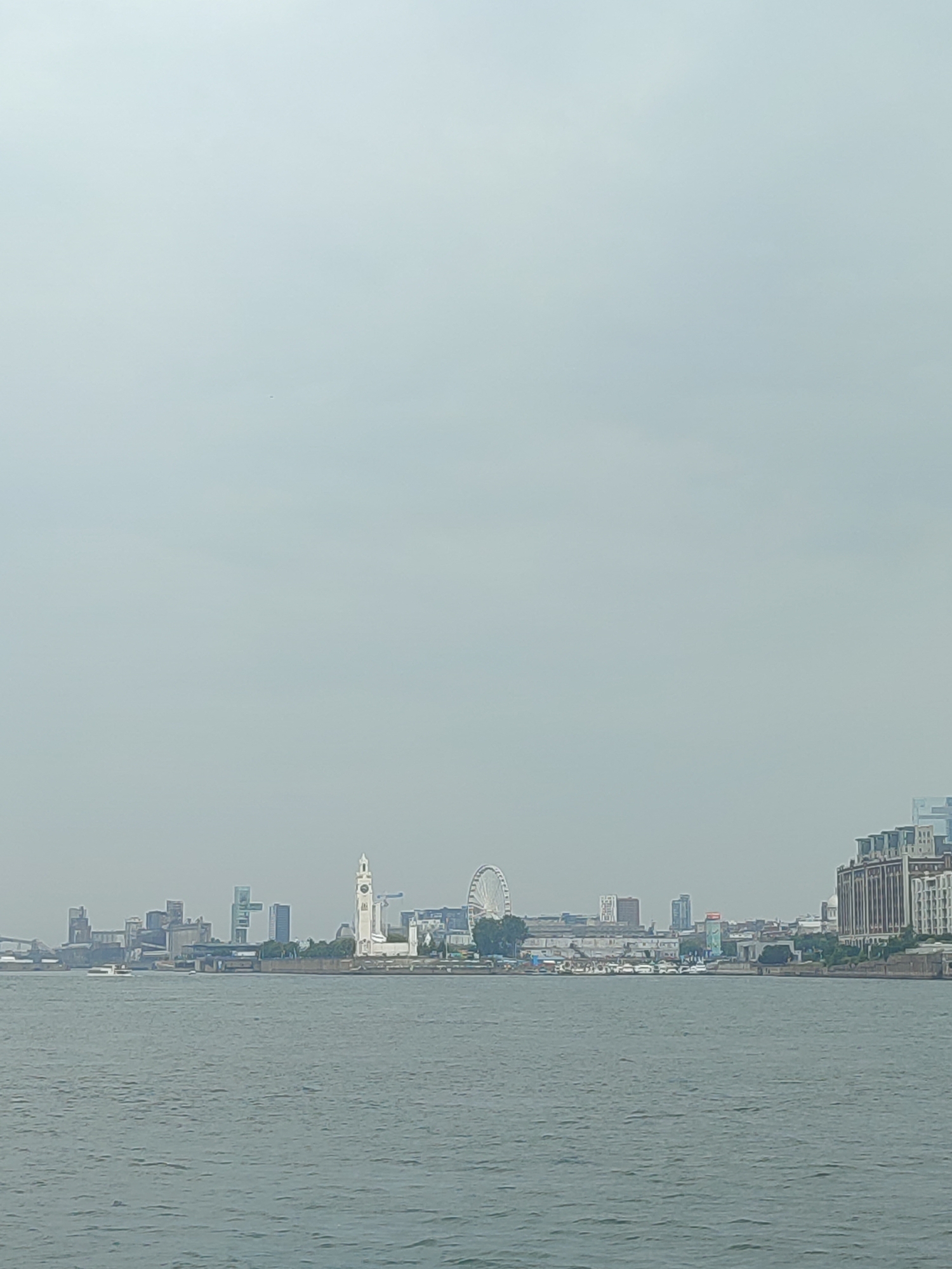 The image captures a waterfront view of Montreal, Canada, under an overcast sky. The foreground is dominated by the rippling surface of the water, reflecting the muted light. In the middle ground, a line of buildings forms the city skyline, with a prominent clock tower standing out. To the right of the clock tower, a Ferris wheel is visible. Further to the right, a cluster of large, multi-story buildings with distinct architectural features lines the waterfront. The sky is a uniform pale gray, suggesting a cloudy or foggy day. No people are discernible in the image, and no specific actions or interactions are depicted. The scene is a static panorama of the city's waterfront infrastructure.