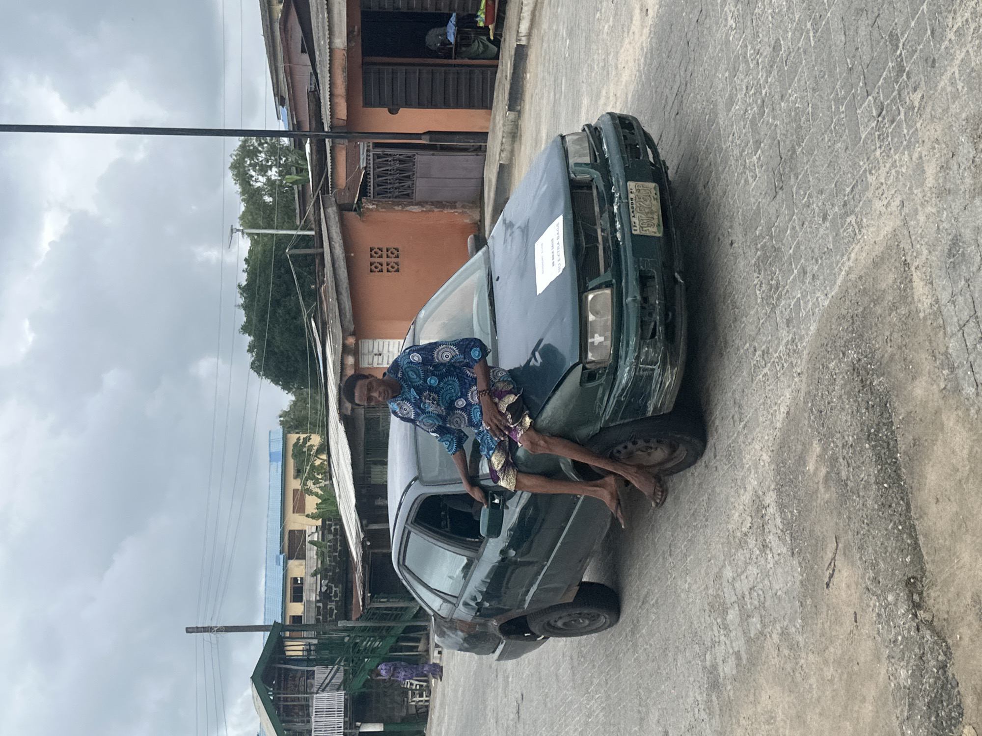 A dark green, weathered sedan with visible damage to its front bumper and paint is parked on a cobblestone street in Badagry, Nigeria. A young man wearing a blue and white patterned shirt and patterned shorts sits casually on the hood of the car, with his legs crossed and one hand resting on the side mirror. His feet are bare, and he wears sandals. A white paper is affixed to the windshield with text that is partially obscured. Behind the car, several buildings with orange and brown facades and corrugated metal roofs are visible. The sky is overcast. In the background, another person in a patterned outfit stands near a structure with a green roof and an external staircase. Power lines are strung overhead. The overall scene depicts a street in a developing urban area.