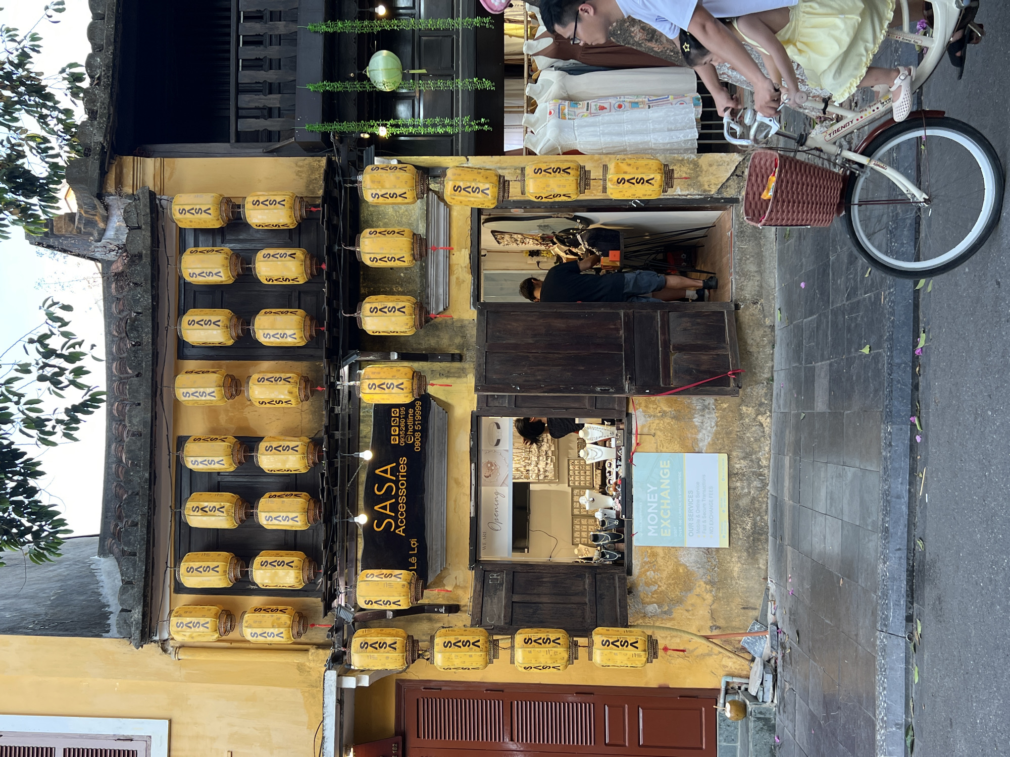 The image depicts a street scene in Hội An, Vietnam, specifically in Hội An Ward. The focal point is a yellow building adorned with numerous hanging yellow lanterns, each bearing the "SASA" logo. Below the lanterns, a shop named "SASA Accessories" is visible with its interior displaying jewelry and accessories. A "Money Exchange" sign is posted outside the shop.
On the right side of the image, a person is riding a bicycle with a young child seated in front. The child is wearing a yellow dress and a star-shaped hair clip. The bicycle has a wicker basket on the front. The street is paved with tiles and asphalt, and there are fallen flower petals on the ground. The building features traditional Vietnamese architecture with dark wooden shutters and balconies. Green vines hang from the upper levels of the building on the right.