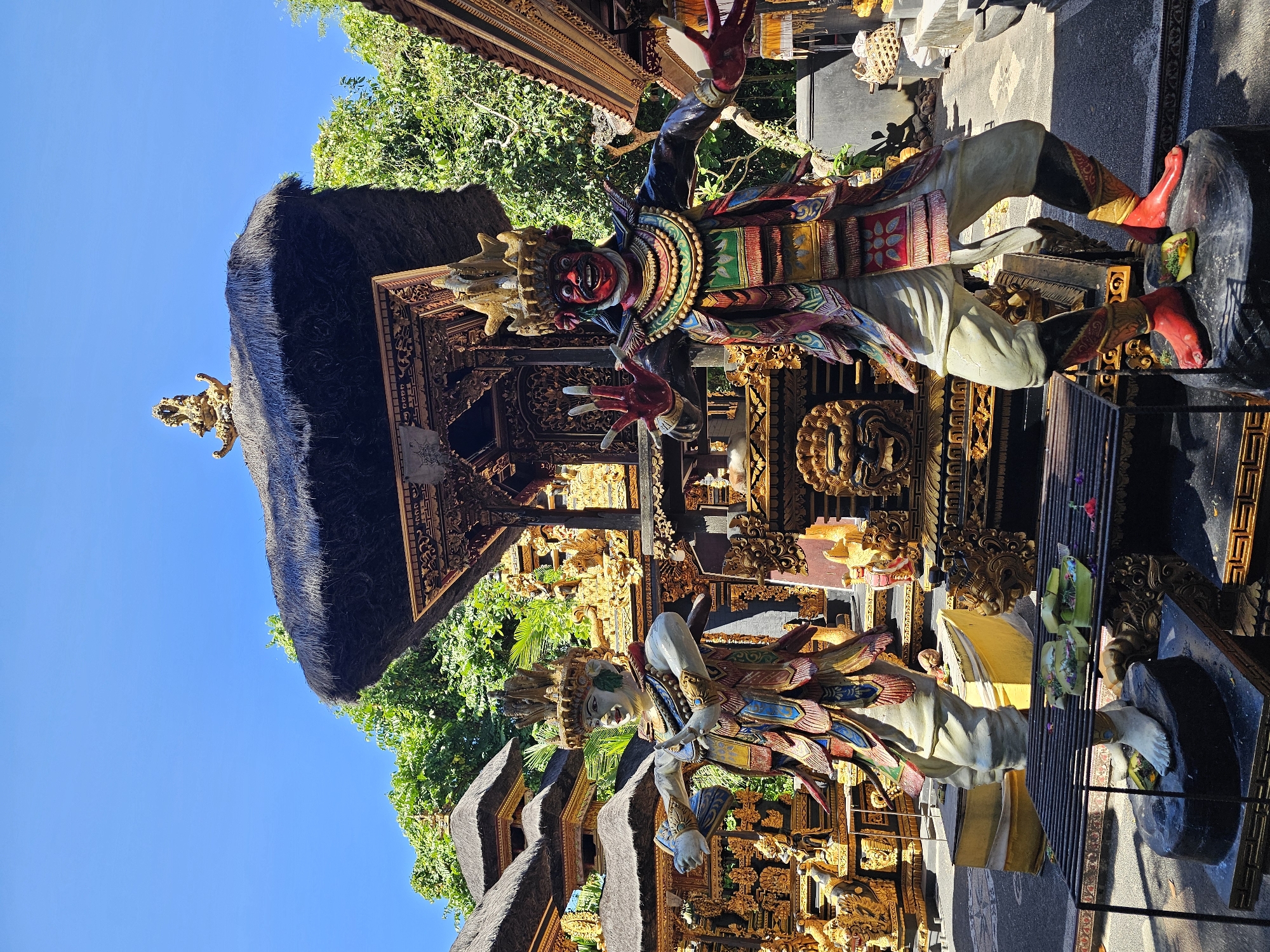 The image shows a temple in Kuta Selatan, Indonesia. Two statues stand in front of a building with intricate carvings and a thatched roof. The statues are brightly painted and appear to depict mythological figures. The upper statue has a red face and outstretched hands. The lower statue has a white face and its arms are bent. The environment includes trees and a clear blue sky. There is a black metal shelf in front of the lower statue with two yellow items on it, in front of a grey pavement.