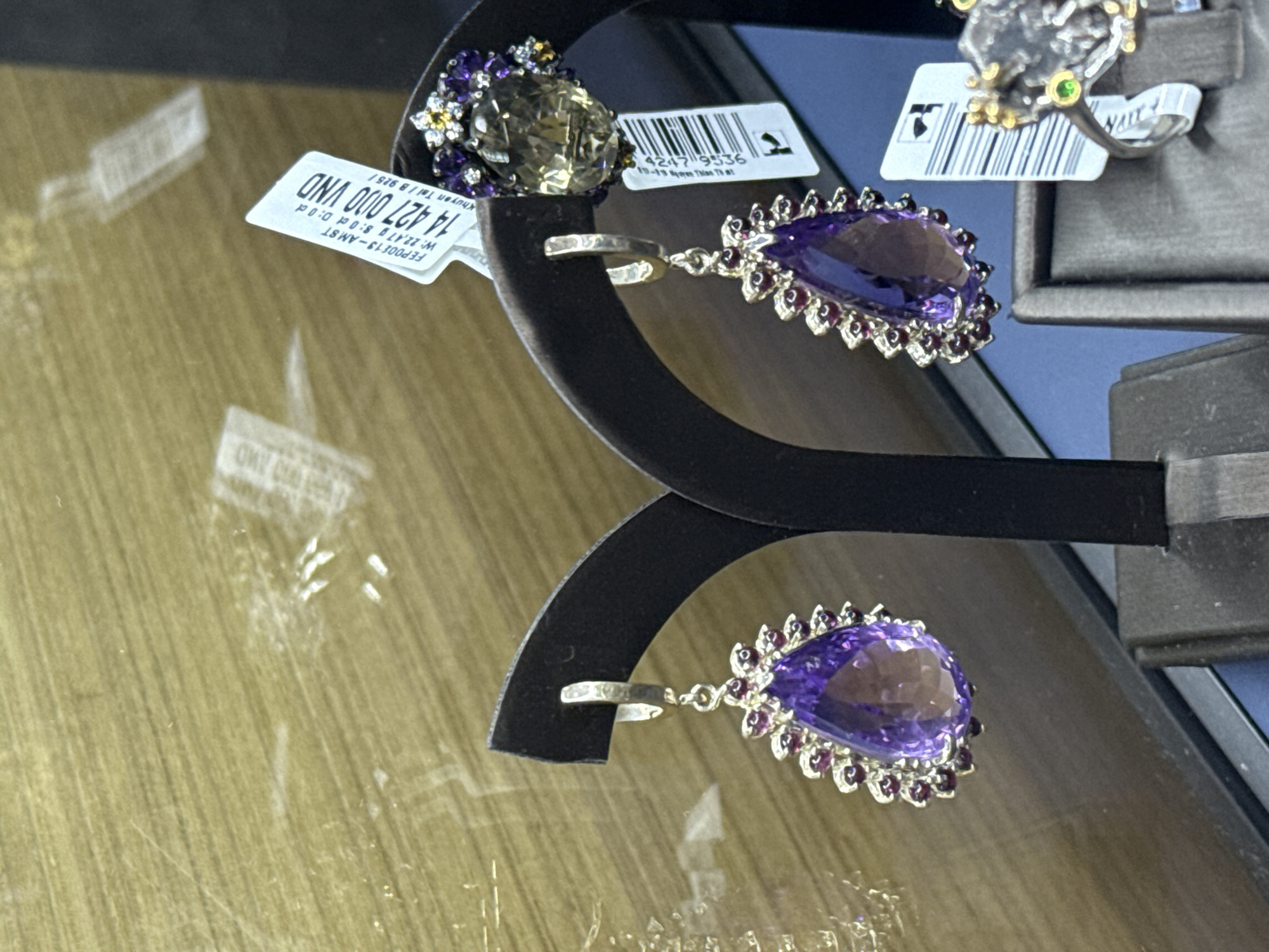 Here is a detailed description of the image:
**Scene:** A close-up shot of jewelry on display in a store.
**Objects:**
- A pair of earrings with large, pear-shaped, purple stones surrounded by smaller stones.
- A ring with a brownish-yellow stone surrounded by purple and white stones.
- Price tags with barcodes and monetary values written in Vietnamese currency (VND), specifically 14,427,000 VND.
- Black display stands holding the jewelry.
**Environment:**
- The jewelry is displayed on what appears to be a wooden or faux-wood surface.
- A piece of paper is visible underneath the jewelry.
**Labels (from price tags):**
- "FEPO0E13-AM ST"
- "W: 22,47 g 8:0 ct D: 0 d"
- "Khuyên Tai / B 925/"
- "13-19 Nam Thim That"
- "4247 9536"
**Location Clues:**
- The currency is VND, indicating Vietnam.
- One of the price tag labels says "13-19 Nam Thim That", which may be a local street or address. If that is a street name, then "Nạm Thím That" might be near Nha Trang Ward, Vietnam.