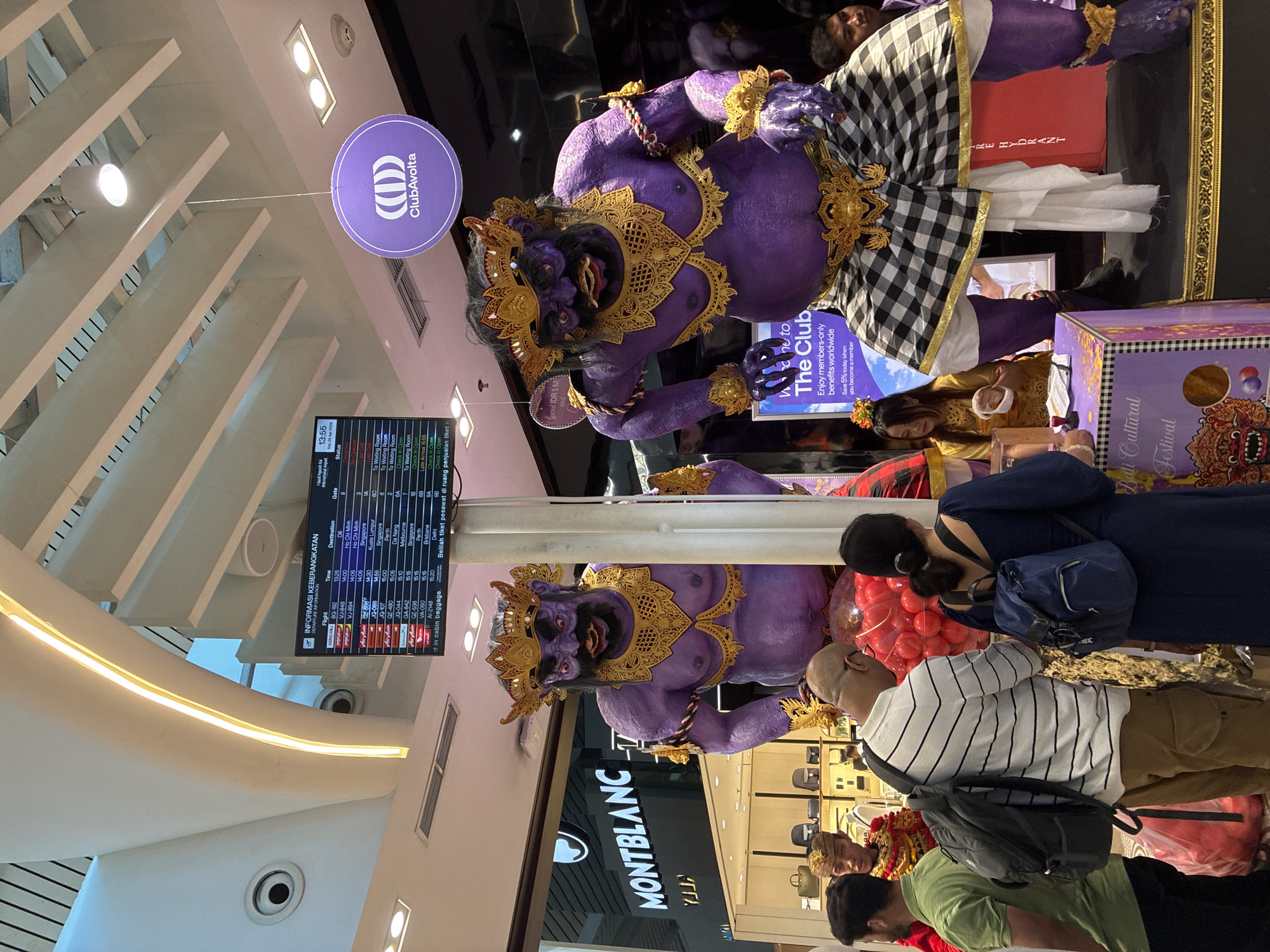 This image shows a scene inside an airport, likely in Kuta, Indonesia, featuring a large, purple statue of a Balinese Barong figure. There are two statues of Barong. The statue is adorned with gold-colored ornamentation and a checkered textile around its waist. A “ClubAvolta” sign hangs near the statue. An airport departure board displays flight information in the background. People are looking at the Barong statues. Some are facing the statues, indicating that they are paying attention to the statues. A "MONTBLANC" store can be seen behind the lower statue. There is a sign that says, "Welcome to the Club".
