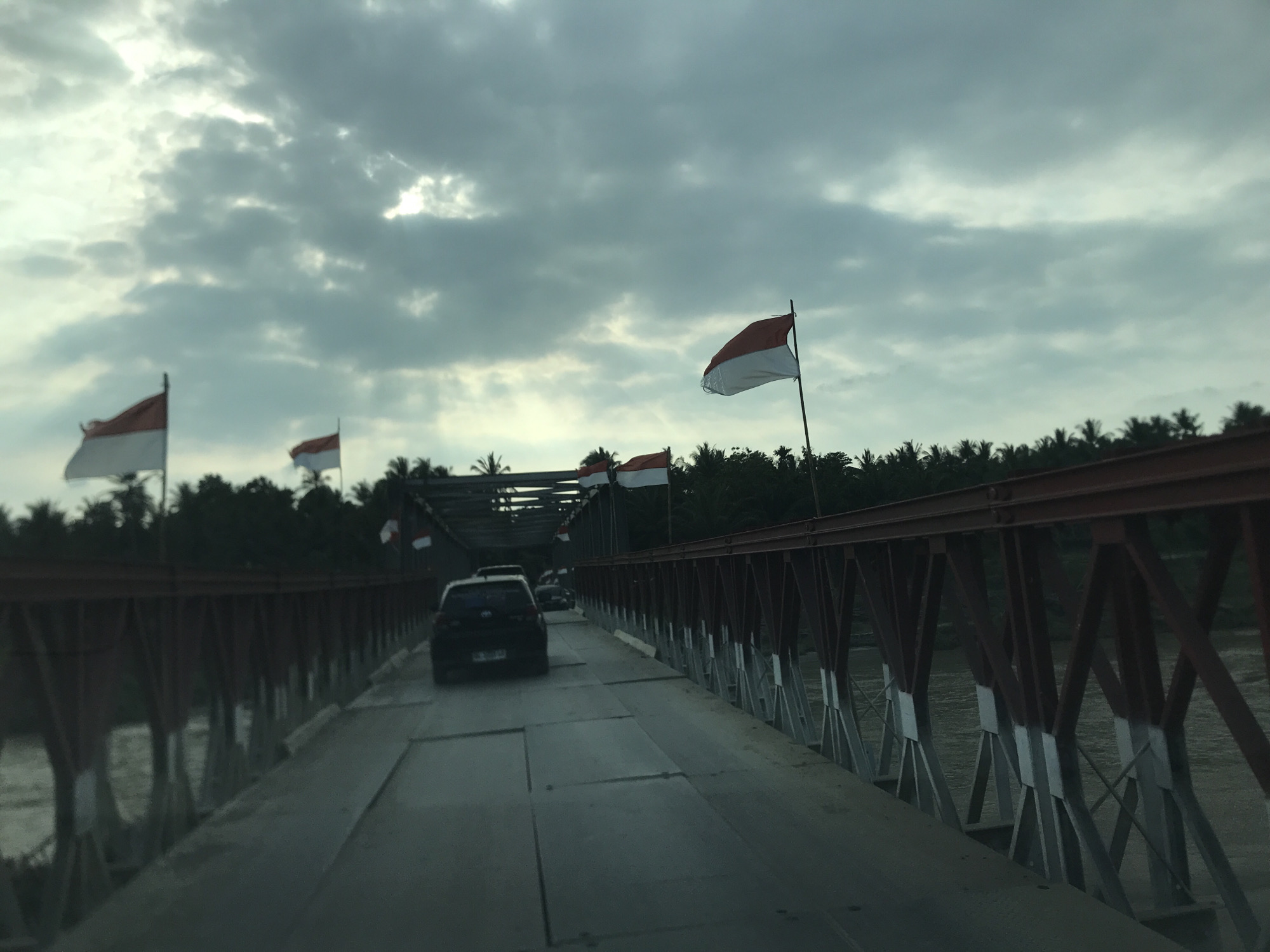 The image shows a road bridge in Awegeutahpaya, Indonesia. Several cars are driving along the bridge. Red and white Indonesian flags are placed on the sides of the bridge. Below the bridge, there is a river. The sky is overcast. Trees line the riverbanks in the background. The bridge appears to be made of steel and painted with a combination of dark red and silver/grey.