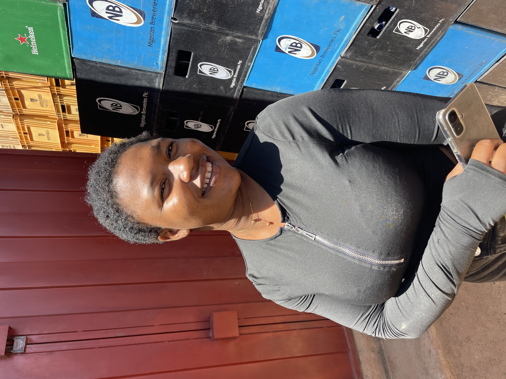 The image depicts a smiling woman with short hair, wearing a black long-sleeved top with a zipper, and holding a mobile phone. In the background, there is a stack of beer crates with logos of different breweries including Nigerian Breweries Plc, Heineken, and Guinness. The crates are various colors including blue, black, green, and yellow. A portion of a red or maroon wall is also visible in the lower left corner. Based on the brewery logos, the location is likely Gura Top, Nigeria.