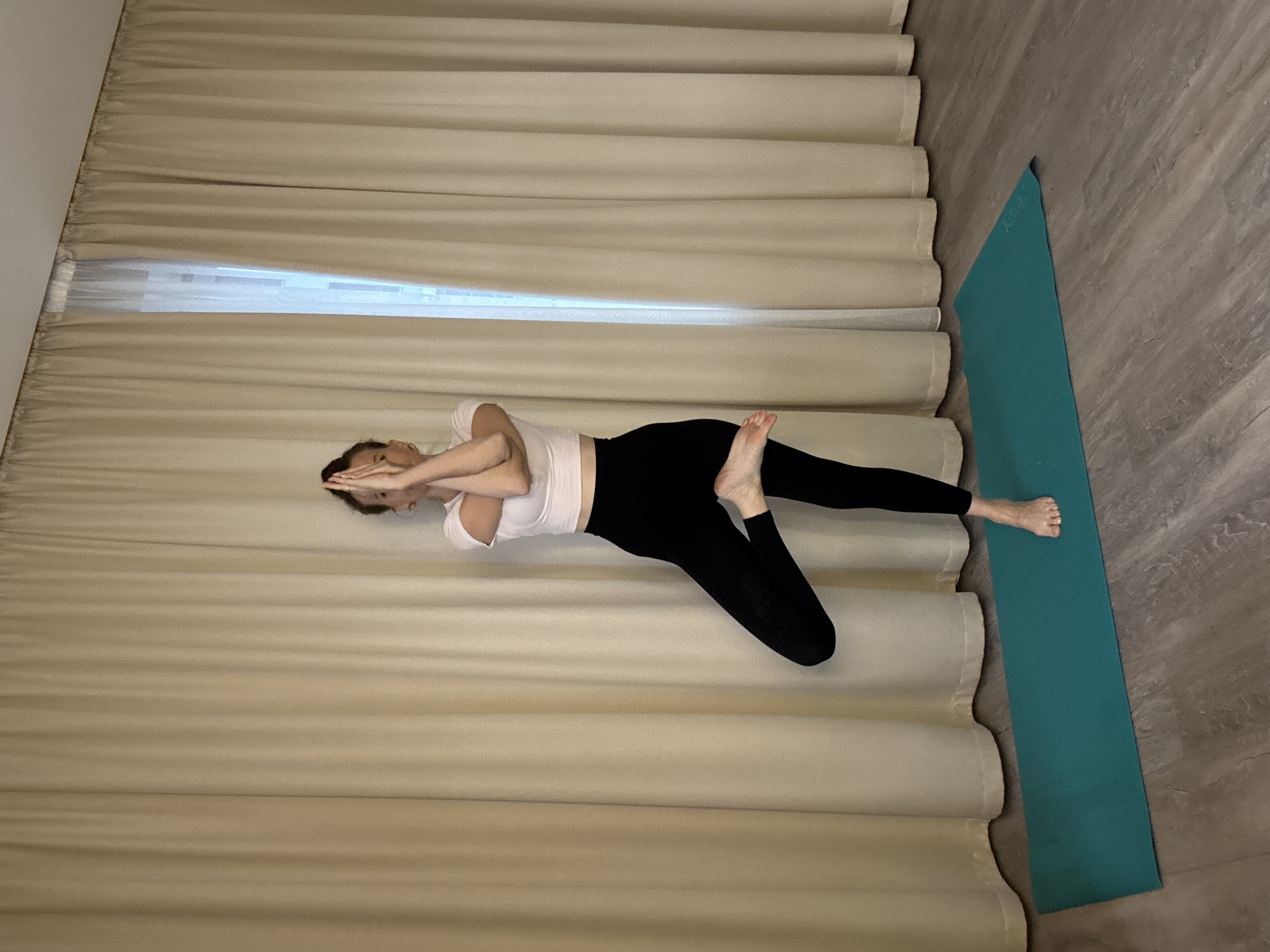 A woman is practicing yoga in a room with light-colored curtains. She is standing on a teal yoga mat on a wooden floor. The woman is wearing a white crop top and black leggings. She is in a tree pose, with her right leg bent and her right foot resting on her left thigh. Her arms are crossed in front of her chest with her hands in a prayer position. The room is dimly lit, with light coming from a window behind the curtains. The overall scene appears to be a home or a yoga studio. There is no information available about the specific location in Moscow, Russia.