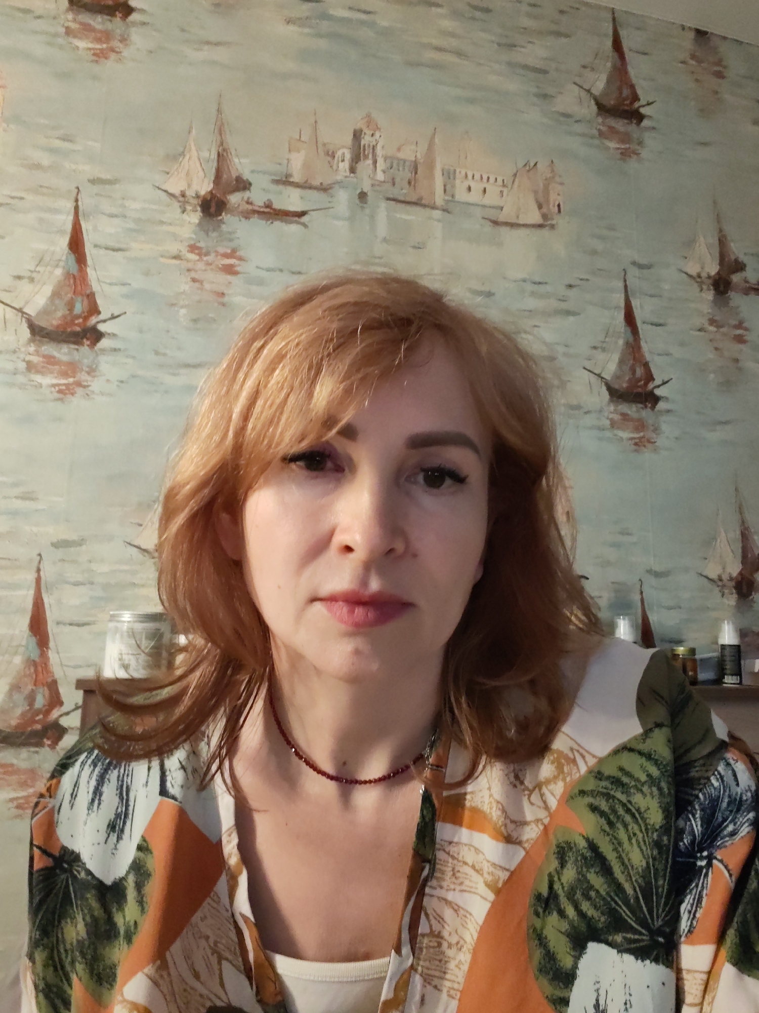 An adult female with light reddish-brown hair and fair skin is depicted from the chest up, centered and facing the camera with a neutral expression. She wears a dark red beaded choker necklace and an open-collar shirt featuring large green, orange, and light brown leaf patterns on a white background, over a white undershirt. Her makeup includes defined eyebrows, winged eyeliner, and pink lipstick. The background is an interior wall adorned with wallpaper illustrating a maritime scene, showing numerous sailboats with reddish sails on light blue water, and faint light-colored buildings in the distance. On a surface to the viewer's left, a light-colored jar with a dark lid is partially visible. To the viewer's right, multiple bottles and containers are partially obscured on another surface. This indoor environment does not provide visual cues to confirm its specific location within the city of Chaikovsky, Russia.