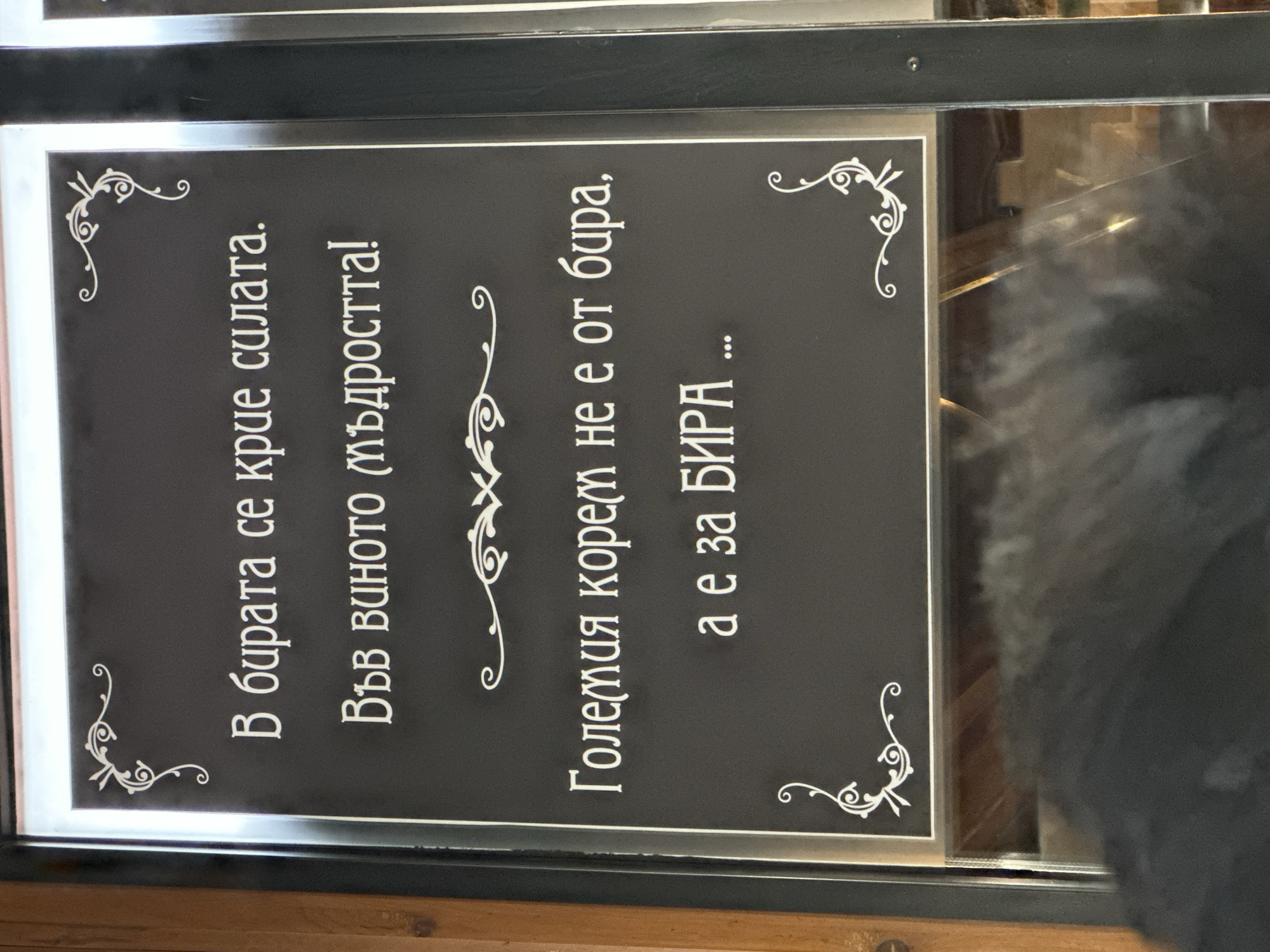 The image shows a framed sign with white text on a black background, likely displayed in a bar or restaurant. The text is in Bulgarian and contains humorous sayings related to beer and wisdom. A corner of the sign includes decorative flourishes. The sign seems to be displayed behind a glass panel or window, and a blurred figure is visible in the reflection. The location can be deduced from the text being in Bulgarian, and this is consistent with the request of Sofia, Bulgaria.