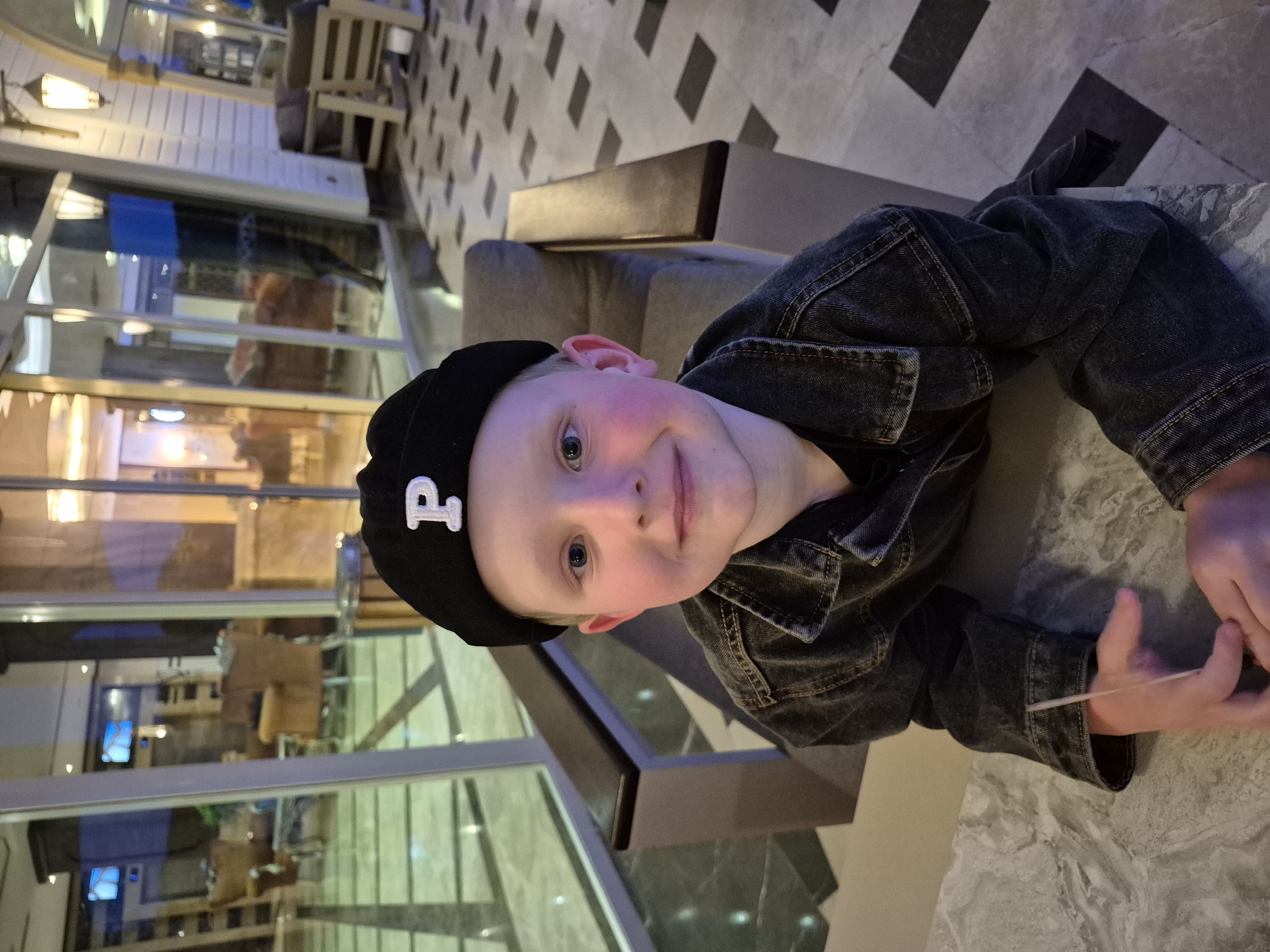A young boy wearing a black cap with a white "P" sits at a table in an indoor location. He wears a dark denim jacket. He is holding a thin, light-colored object between his fingers. The background suggests an interior with glass partitions reflecting the environment, possibly a hotel or similar establishment in the city of Alanya, Turkey. The flooring is patterned, and there is furniture visible in the background, indicating a public seating area.