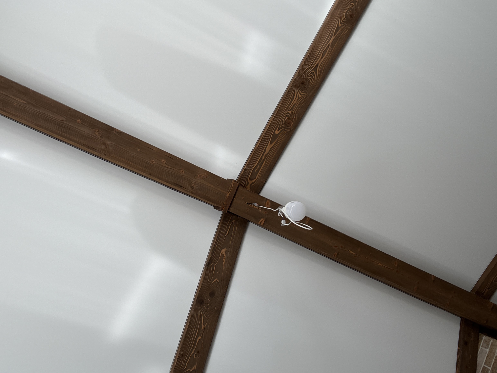 The image shows a white ceiling with exposed dark wooden beams forming an X shape. A small, round white light fixture is hanging from one of the beams by a wire. The lighting suggests the room is illuminated, casting shadows on the ceiling. The beams have a visible wood grain and knots, indicating they are natural wood. Part of a brick wall is visible in the bottom left corner of the image. The overall scene depicts an interior architectural detail, possibly in a modern or rustic setting. The location in Georgiyevsk, Russia is not visually evident from the image itself.