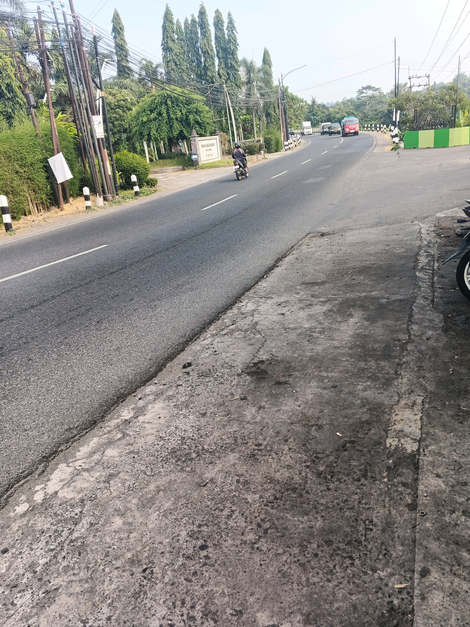 The image shows a street scene in Ungaran, Indonesia, with a road curving to the right. A motorcycle rider is moving away from the camera on the left side of the road. Along the left side of the road, there are numerous utility poles, bamboo plants, and some bushes. A sign that reads "NELVA BALMONIK" is visible behind the poles and bushes. On the right side of the road, there is a paved walkway with a concrete texture. The background features tall trees and a clear sky. In the distance, several vehicles are driving on the road, including a red truck, a white van, and other cars. The overall environment appears to be a suburban or semi-rural area.