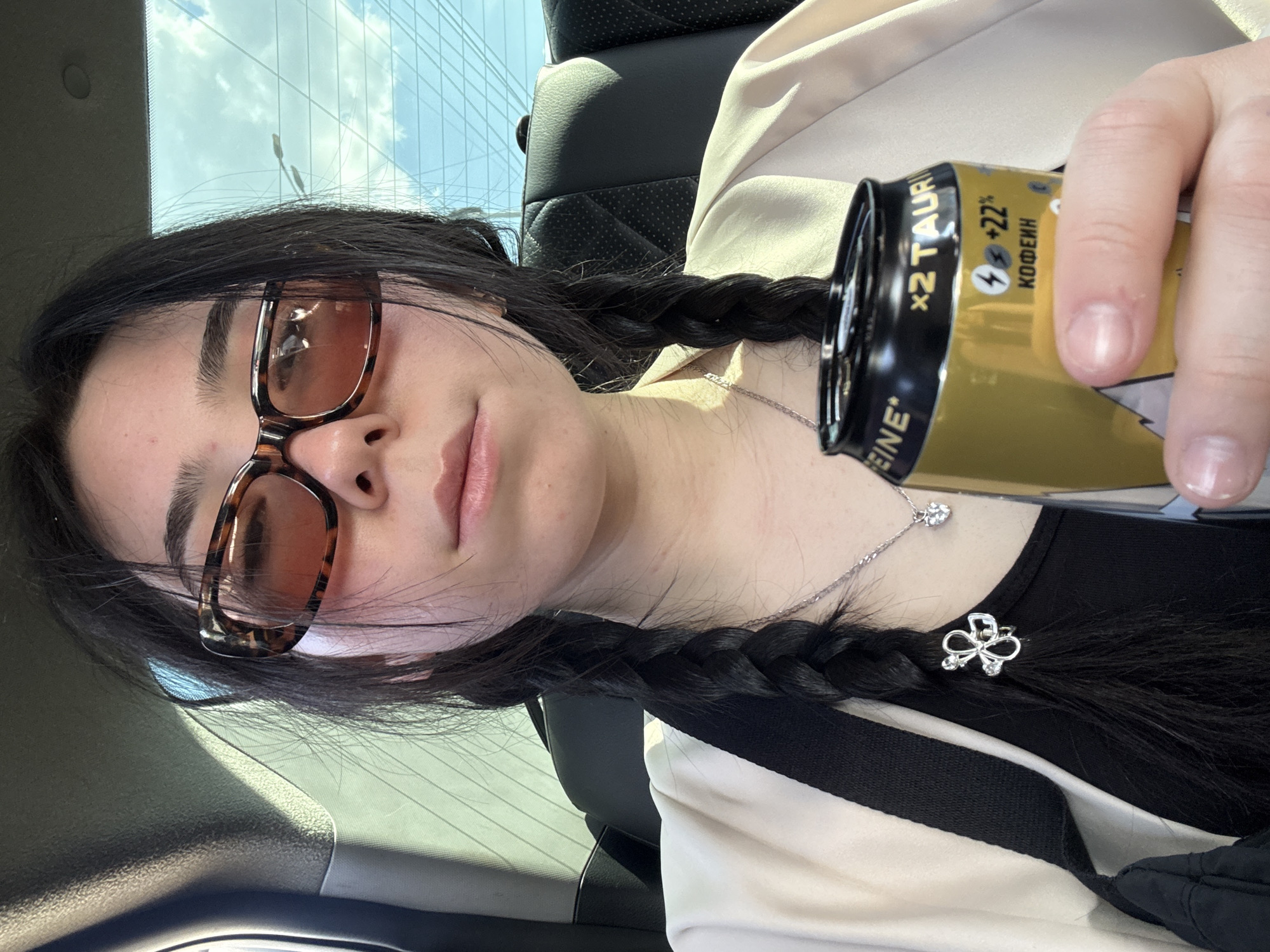 A person with dark hair braided in pigtails, wearing brown tortoiseshell sunglasses, is holding a yellow energy drink can. The can has black writing in Cyrillic and symbols. The person is wearing a white blazer over a black top and has a silver necklace with a small clover pendant. The background shows the interior of a car with a view of a building through the windshield. The setting is likely during the daytime. The location may be Izhevsk, Russia, but there is no clear information to confirm that.