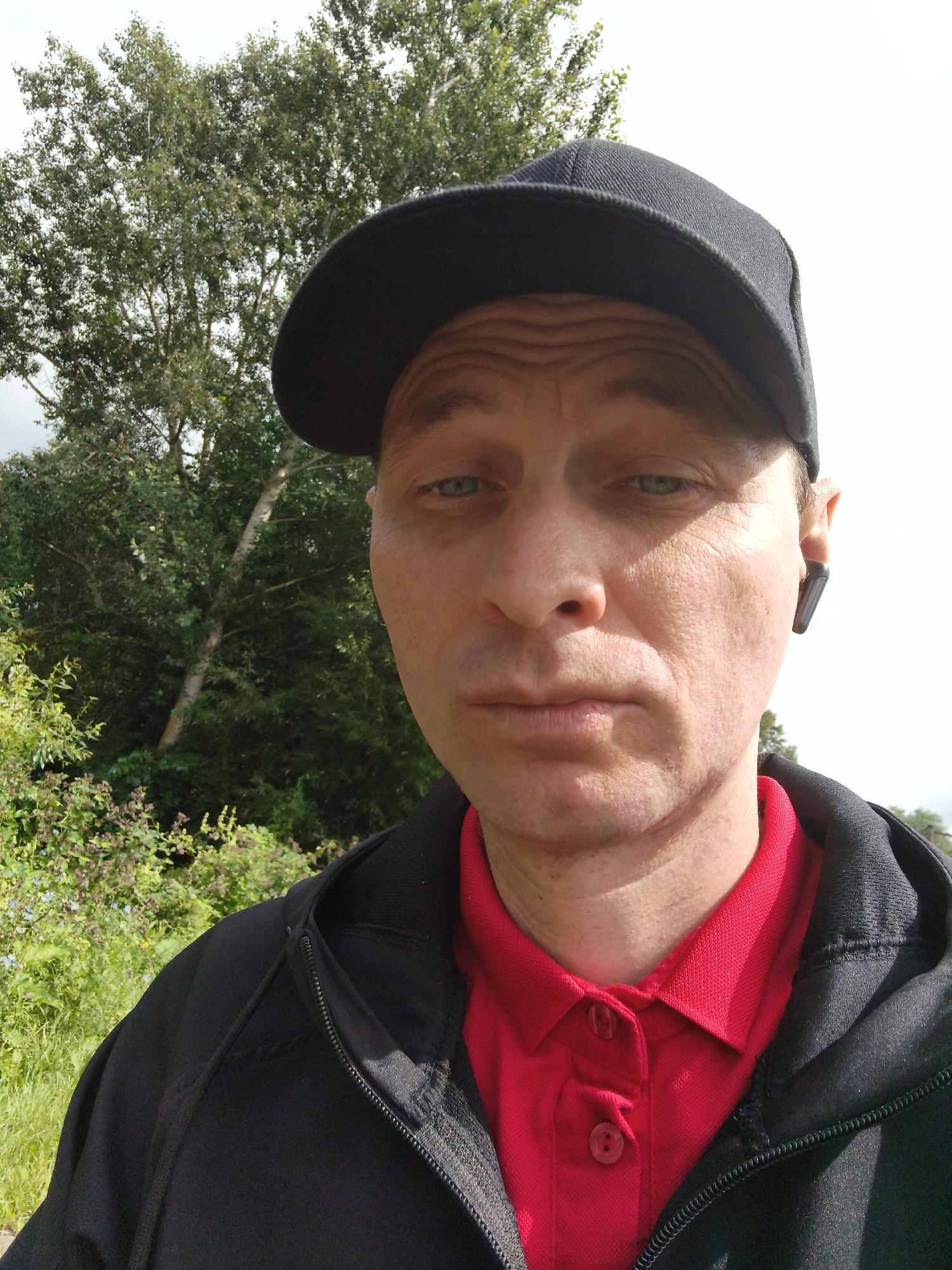 A close-up, over-the-shoulder selfie shows a light-skinned man wearing a black baseball cap, a black zip-up hoodie, and a red polo shirt. He is outdoors, with a dense green treeline and a pale sky in the background. He has short hair, visible stubble, and is wearing a black wireless earbud in his right ear. The man's expression is neutral, and his eyes are open. His face is angled slightly towards the camera, and his lips are closed. The lighting suggests it is daytime, possibly overcast. The man appears to be in a park or natural setting.
