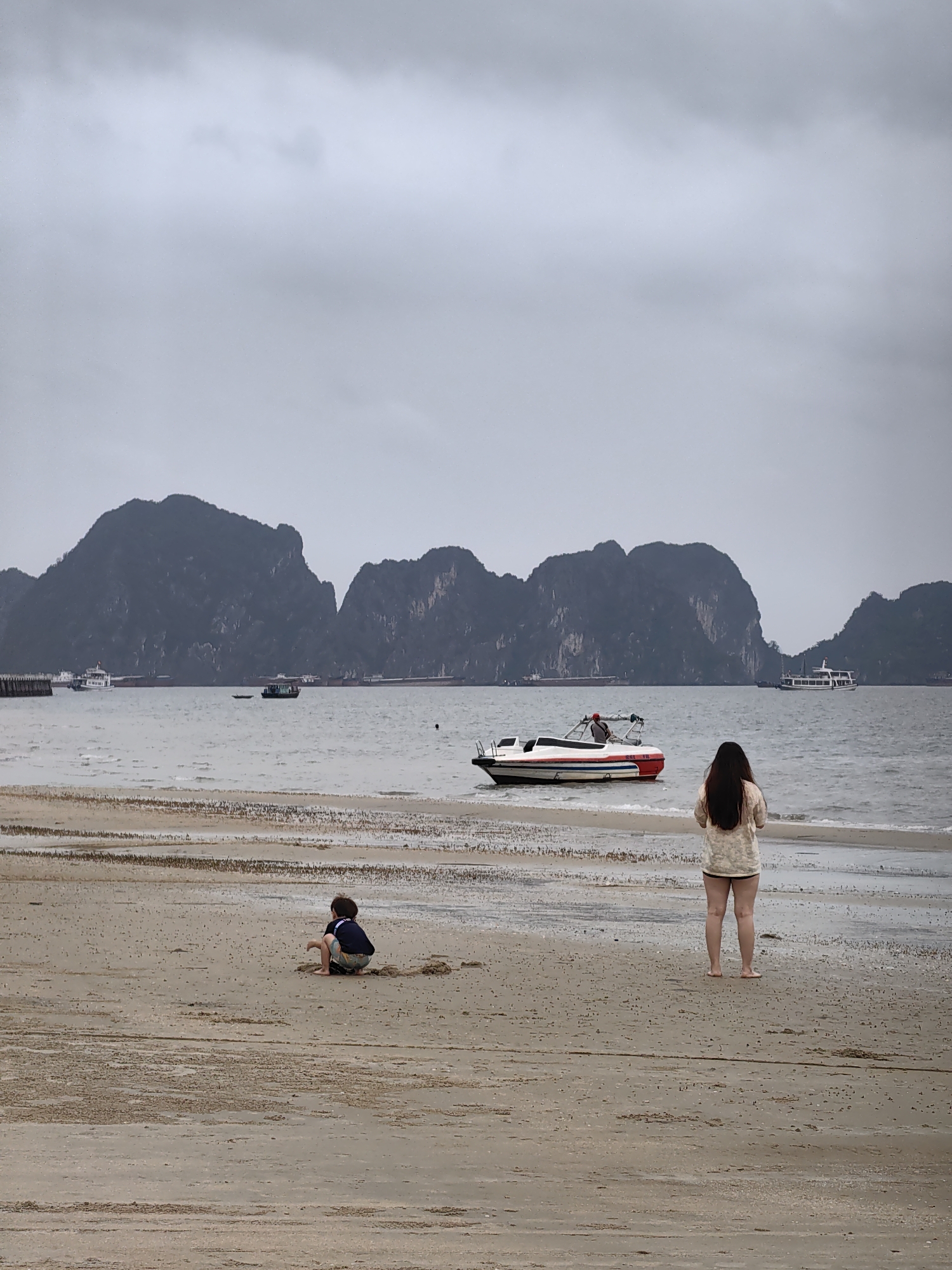 This full-frame, eye-level photograph captures a serene, overcast day on a beach in Bai Chay Ward, Vietnam, showcasing the iconic karst limestone islands of the Ha Long Bay region in the background.
The sky is uniformly gray and cloudy, extending over a choppy, dark gray-blue sea. In the distance, several large, rugged, dark green and gray mountains rise majestically from the water. Closer to the mountains, several boats are visible, including larger passenger vessels and smaller working boats, and a long dark pier or breakwater juts out from the far left shore.
In the mid-ground, a white and red speedboat, marked "DBV 1B", floats in the water, with a person in a red cap visible on board.
The sandy beach occupies the foreground, showing patterns left by the receding tide and scattered debris. Two figures are present on the beach. On the right, an adult female with long dark hair stands facing away from the camera, looking out towards the sea and mountains. She wears a light-colored top and dark shorts, with bare feet in the sand. To her left, in the lower-mid ground, a child is crouched, seemingly playing in the sand, also facing away from the viewer. The child wears dark shorts and a dark t-shirt.
The overall atmosphere is calm and contemplative, depicting a leisurely day at the beach despite the cloudy weather.