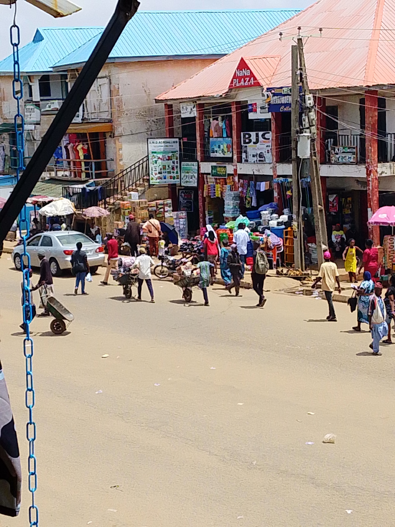 The image depicts a bustling street scene in a marketplace, likely in a city in Nigeria. The foreground is a dusty, paved road with several people walking and engaged in various activities. In the mid-ground, a silver sedan is parked on the left side of the road, and a variety of market stalls are set up with colorful wares displayed. People are seen pushing handcarts laden with goods, while others are browsing or walking. The background features buildings with red and blue roofs, some of which appear to be shops or commercial establishments. A prominent sign reads "NaNa PLAZA". Power lines and a utility pole are visible on the right side of the image. The overall atmosphere is one of lively commerce and daily life in a busy urban environment.