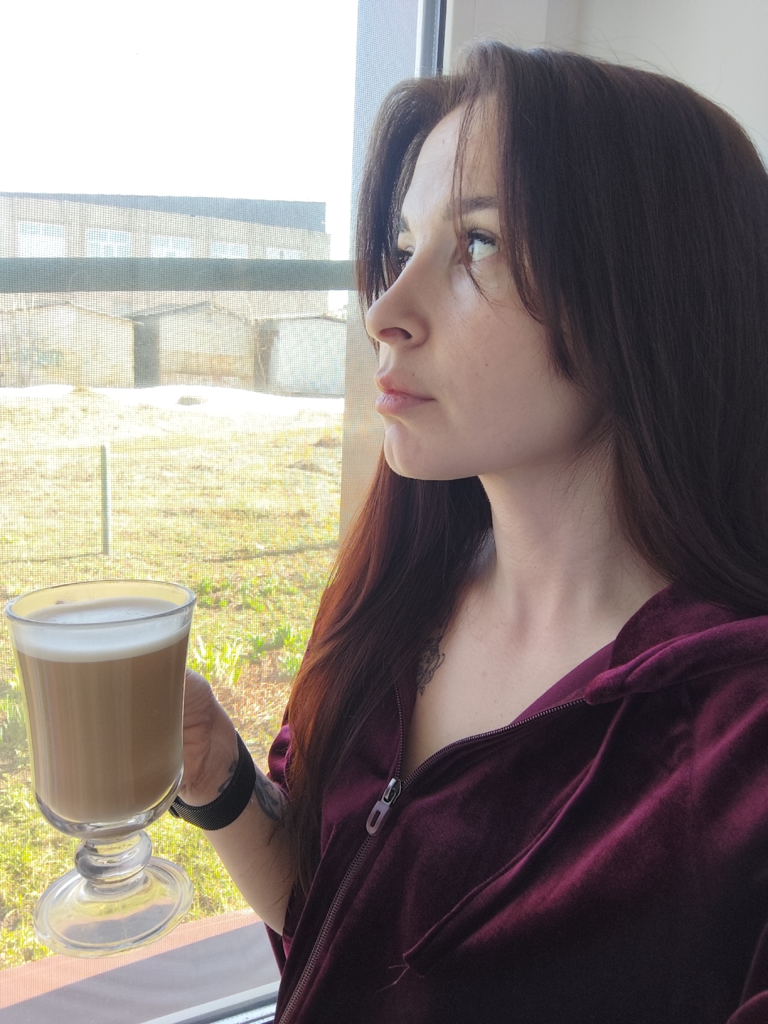 A woman is depicted in a medium shot, holding a glass of latte while gazing out a window. Her attire consists of a maroon, zippered hoodie, with dark hair cascading down her shoulders. A watch adorns her left wrist, and tattoos are visible on her arm and neck. The woman appears to be in Фоки, Russia; a building with several windows is partially visible through the window behind the woman. Beyond the building is a grassy field. The window screen is visible.