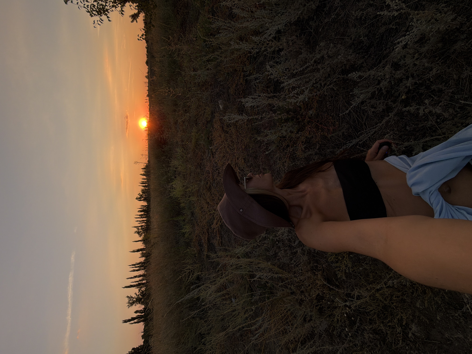 A woman is taking a selfie outdoors during sunset. She is wearing a brown hat and sunglasses. She has long brown hair and is wearing a black bandeau top. A light blue garment is draped over her lower body. The background consists of dry grass and a silhouetted line of trees against an orange and pink sky. The setting appears to be a field outside of Kamyshin, Russia.