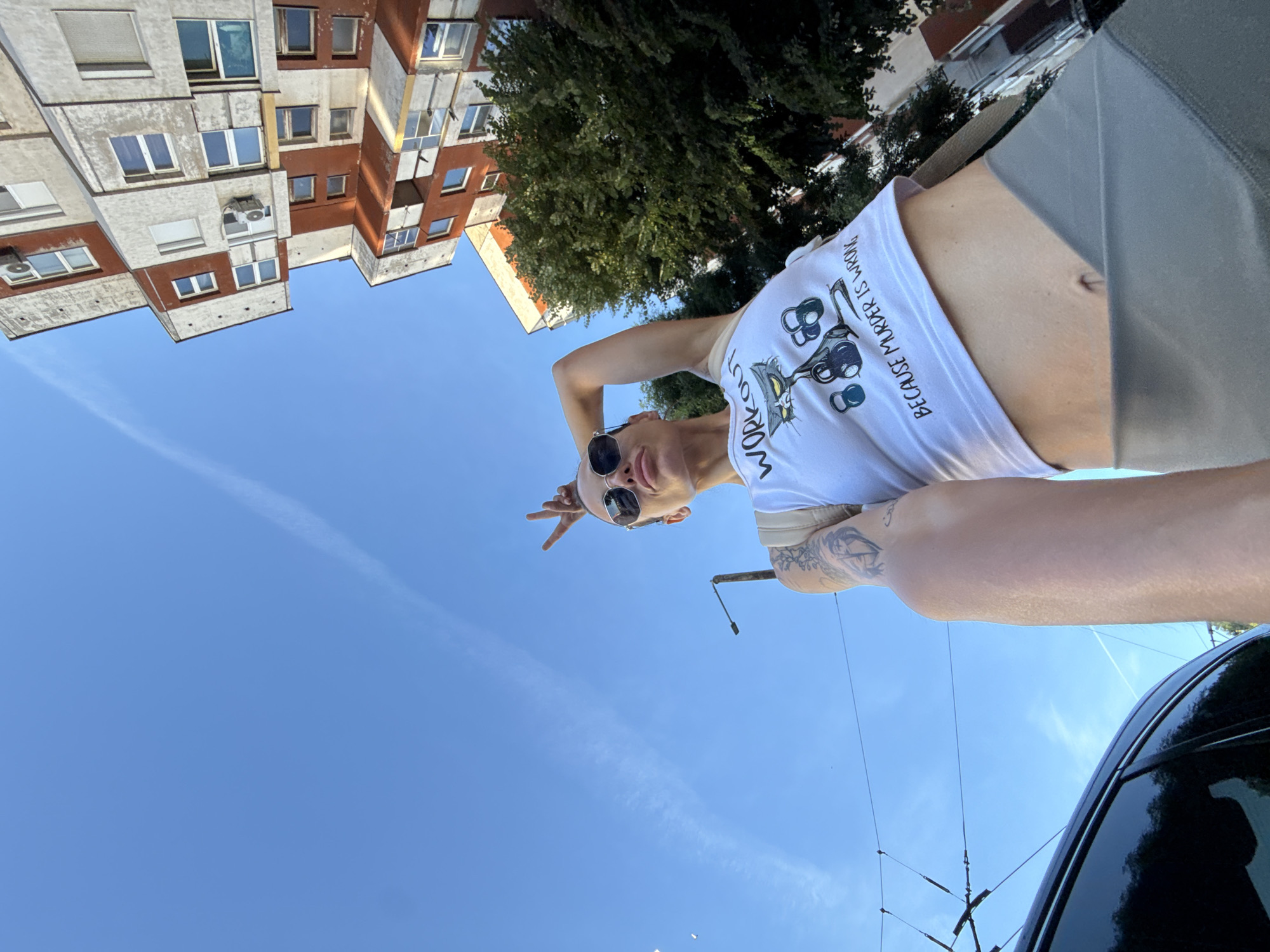 An outdoor, low-angle, full-body selfie depicts a fair-skinned woman in Pleven, Bulgaria. The woman, wearing sunglasses and a white cropped tank top that reads "WORKOUT," has her left arm raised and bent at the elbow, with her fingers in a peace sign gesture near her head. Her right arm is out of frame. She is wearing grey leggings and a beige strap bag is visible over her shoulder. She has tattoos on her left arm. In the background, a multi-story building with alternating orange and white facades and visible air conditioning units stands against a clear blue sky. Green trees partially obscure the building on the right side of the frame. A dark car is visible in the lower left corner. Overhead power lines stretch across the upper portion of the image.
