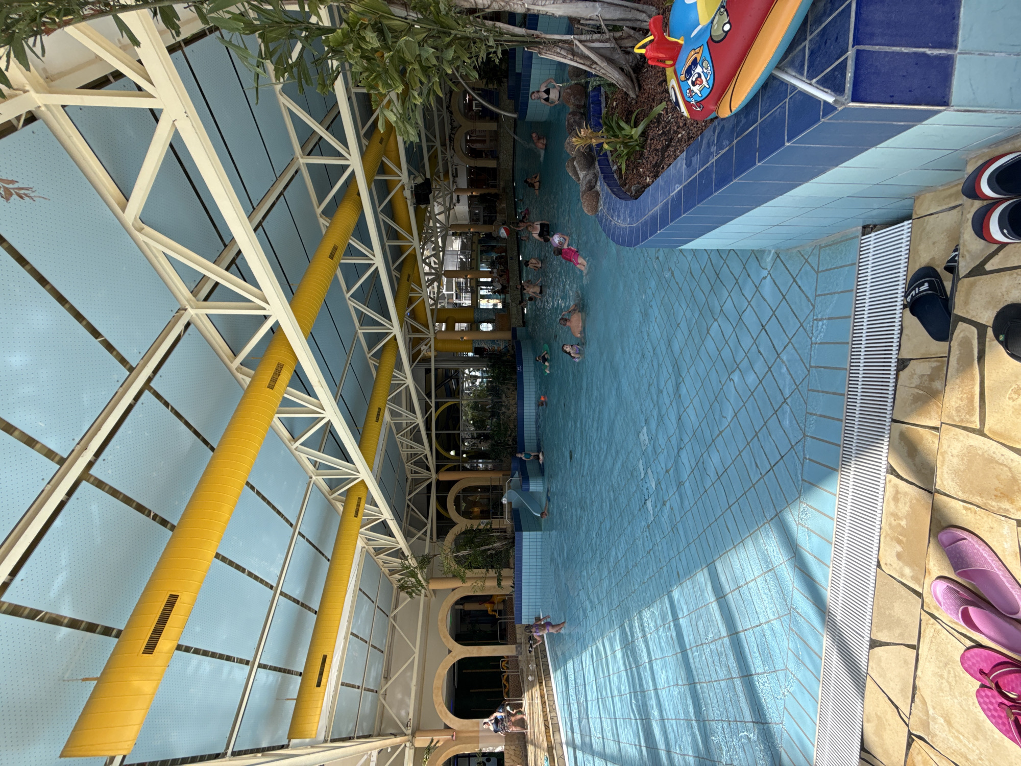 The image depicts an indoor swimming pool facility, possibly part of the "H2O" water park in Rheinbach, Germany, given the context of the location. The pool is large and filled with clear blue water, tiled with small squares. Several people, including adults and children, are visible in the water, engaged in various activities such as swimming, playing, and relaxing. There is a small slide in the pool area. The ceiling of the facility is high and features a light blue translucent material supported by a white metal structure, with large yellow ventilation pipes running along it. The pool is surrounded by a tiled edge and a stone-tiled deck area where several pairs of flip-flops and sandals are placed. Lush green plants and trees are visible both inside and outside the pool area, adding to the atmosphere. The overall environment suggests a recreational and family-friendly setting.