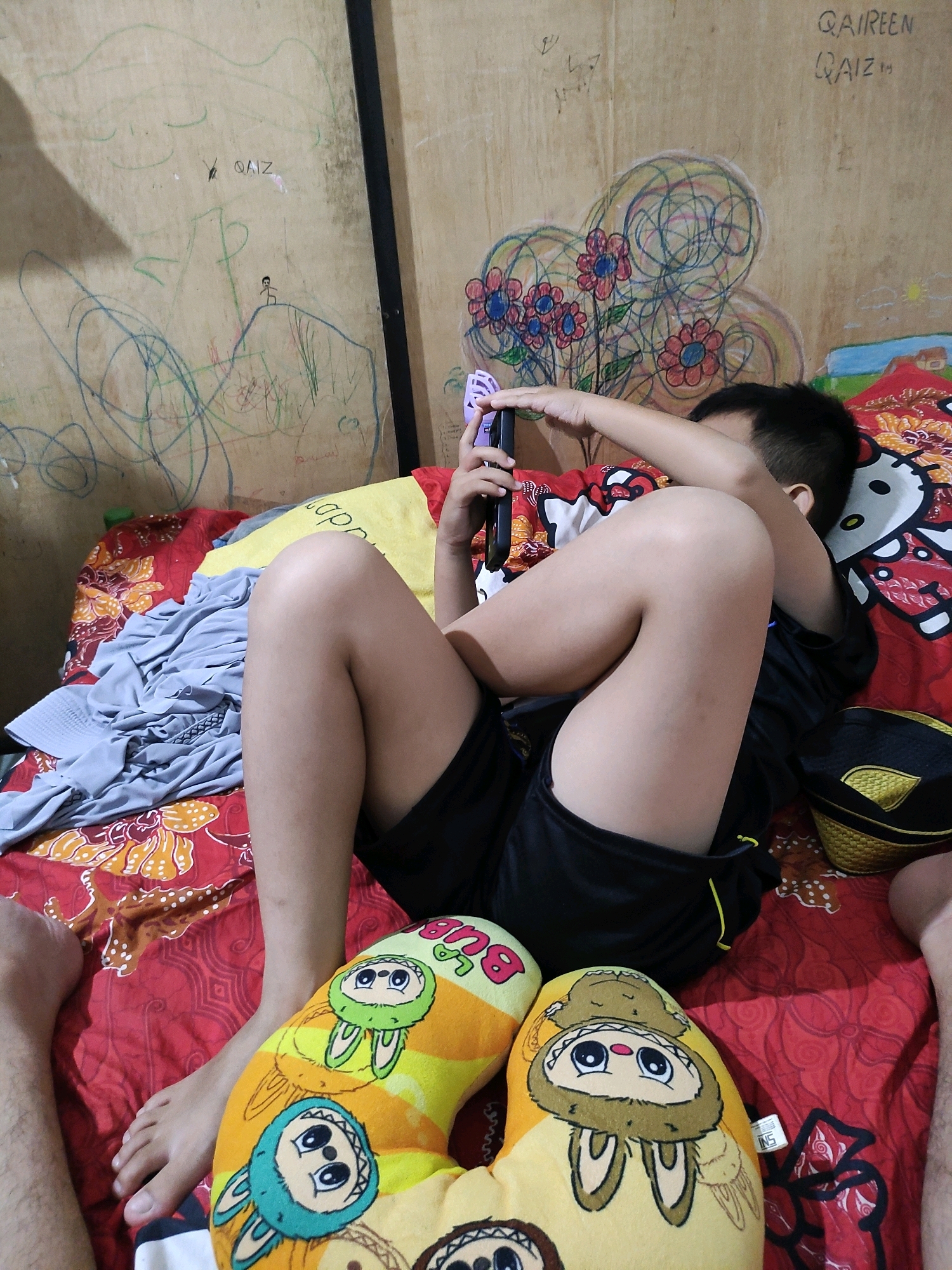 A young boy is lying on a bed, playing on a mobile phone. The bed is covered with a red and white patterned sheet, and a yellow towel is draped over it. There are several pillows on the bed, some with cartoonish designs of animals. The boy is wearing a black t-shirt and black shorts. In the background, the wall is decorated with colorful drawings, possibly made by children. The environment appears to be a home in Bulukumba, Indonesia.