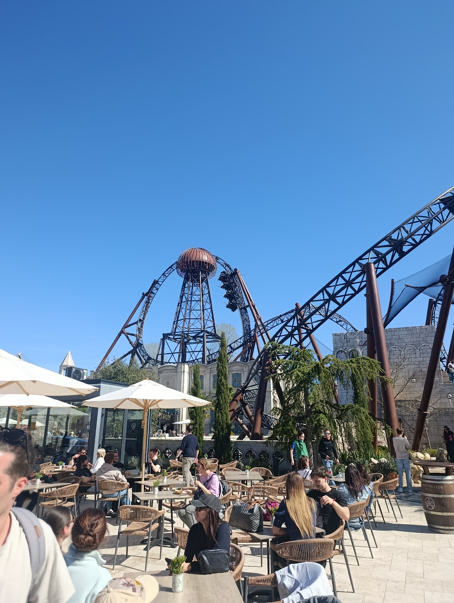 The image shows a sunny outdoor scene at Europa-Park in Rust, Germany. In the foreground, a restaurant patio is visible, with numerous round tables and wicker chairs occupied by people dining and socializing. White parasols provide shade. In the background, a large roller coaster structure dominates the view. The roller coaster has a complex web of dark metal tracks and a prominent spherical element at its peak. Behind the coaster, there is architectural scenery resembling an old European building. The sky is clear and blue. There is a mix of standing and sitting people. A wooden barrel is positioned near the seating area.