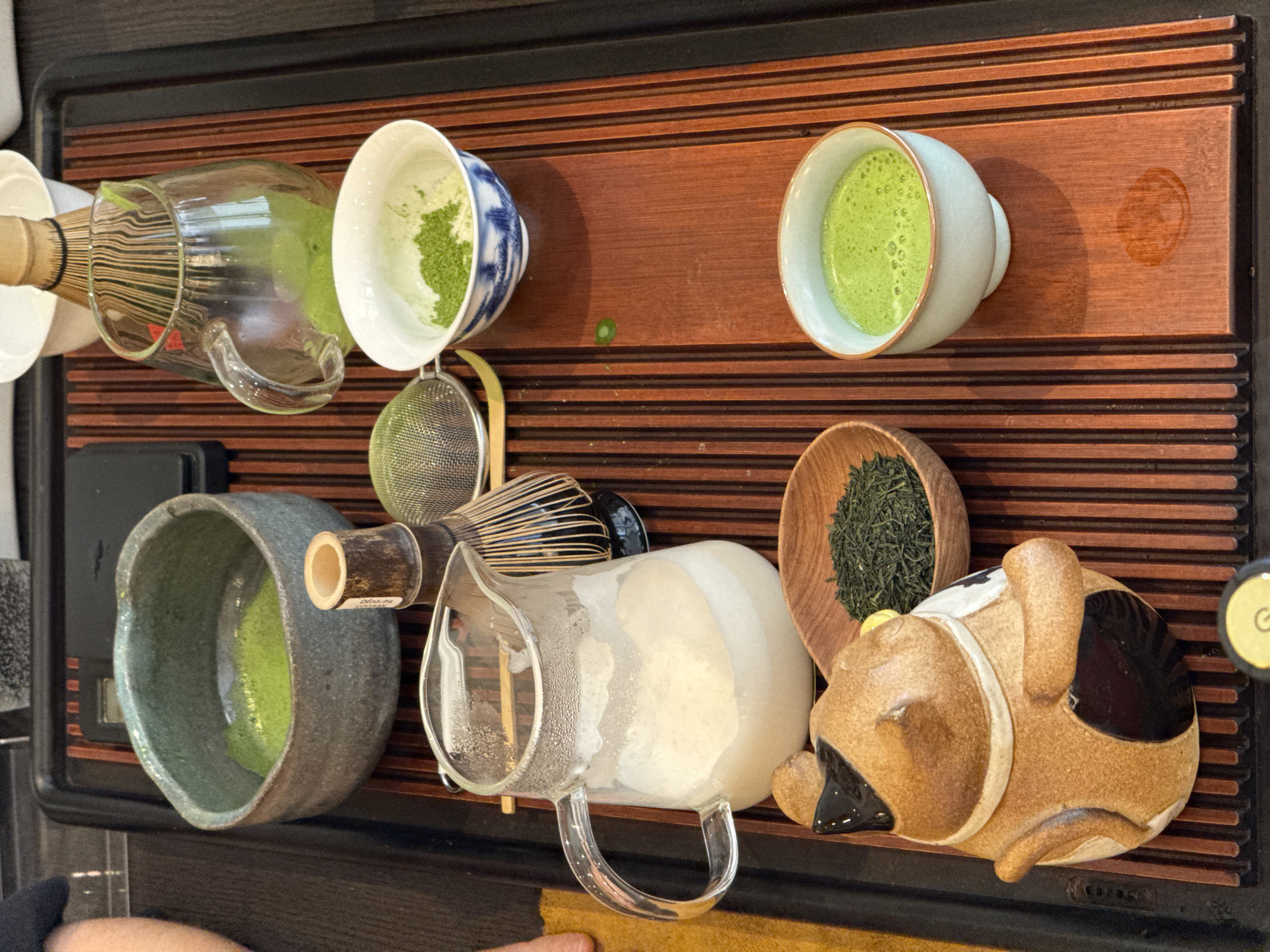 A top-down, close-up shot depicts a matcha tea set on a wooden tray with alternating dark and light brown horizontal stripes. From left to right along the top edge of the image is a container holding a whisk, a glass container, a cup with green powder in it, and a white cup filled with a green liquid. From left to right along the bottom edge is a bowl filled with a green liquid, a whisk on top of a glass container filled with a white liquid, a small wooden bowl of green tea leaves, and a brown animal-shaped object. The photo was most likely taken in the city of Budva, Montenegro.