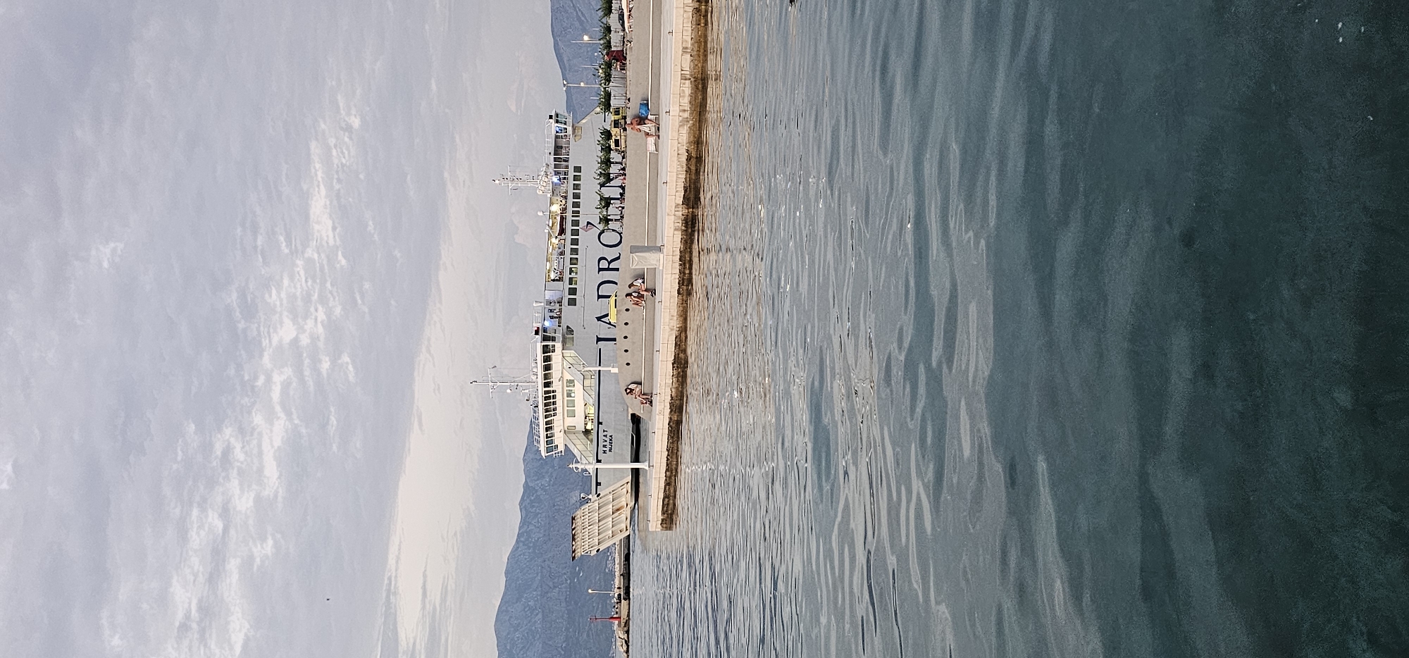In Grad Supetar, Croatia, a wide-angle, vertical photograph captures a ferry docked at a pier. The ferry, white with blue lettering that reads "JADROLINIJA," is positioned on the left side of the image. Its ramp is lowered. The pier, a light-colored concrete structure, extends from the bottom right corner of the frame and curves towards the ferry. Several people are scattered along the pier, some sitting, others standing or walking. In the foreground, the calm, dark blue water of the harbor reflects the sky and the structures. In the background, a distant mountain range is visible under a cloudy, overcast sky. A red lighthouse stands on a small island to the far left. The scene appears to be set during the day, possibly late afternoon or early morning due to the soft lighting.
