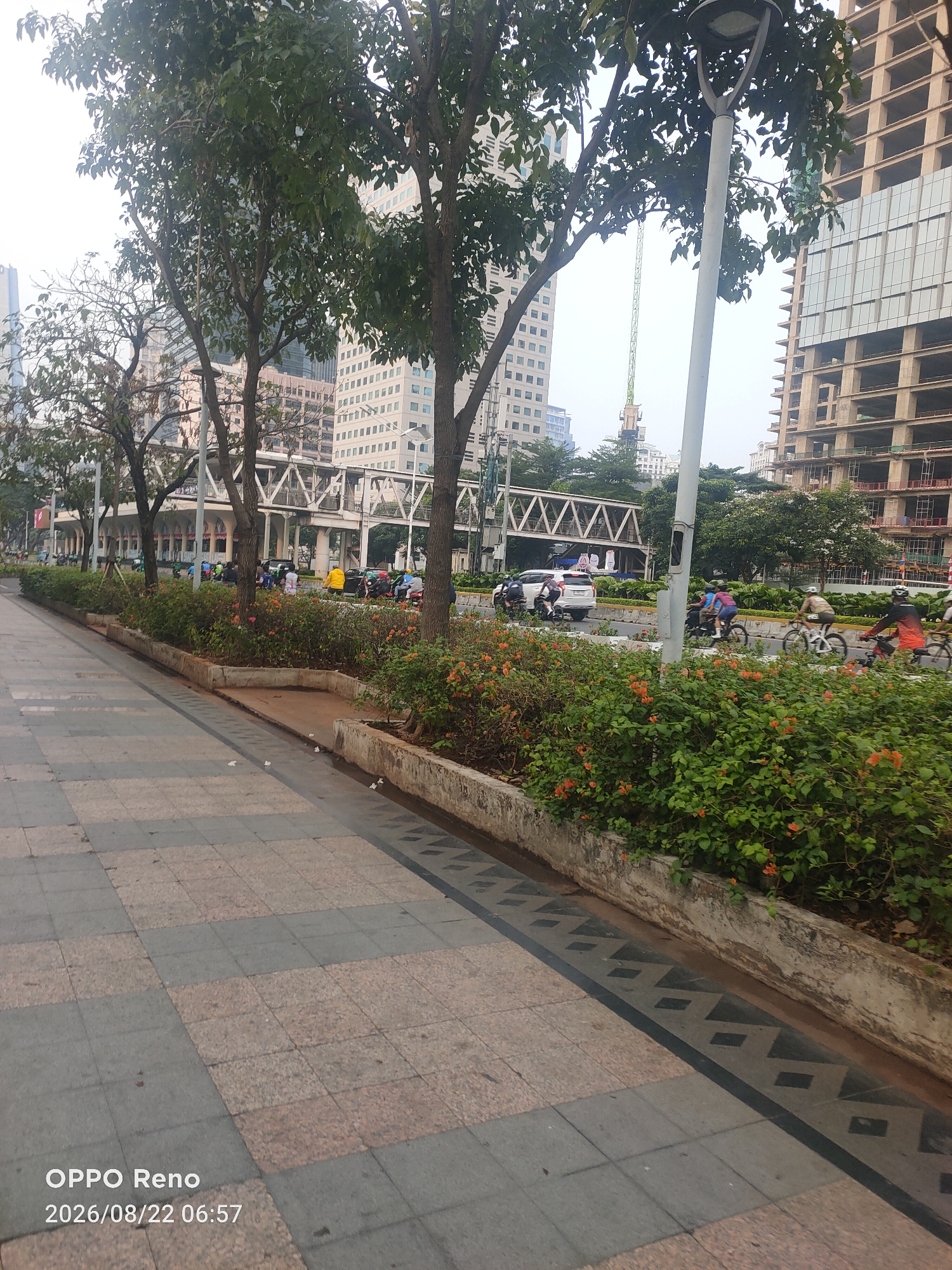 In an urban environment, a paved walkway is visible in the foreground, with planters containing lush green foliage and orange flowers lining the edge. Beyond the planters, a road with multiple lanes is present, featuring traffic including cars, motorcycles, and bicycles. Several individuals are seen riding bicycles on the road. In the background, tall buildings, some appearing to be under construction, dominate the skyline. A pedestrian bridge spans across the road. Trees line the walkway and the road. The overall scene suggests a busy city street with ongoing development.