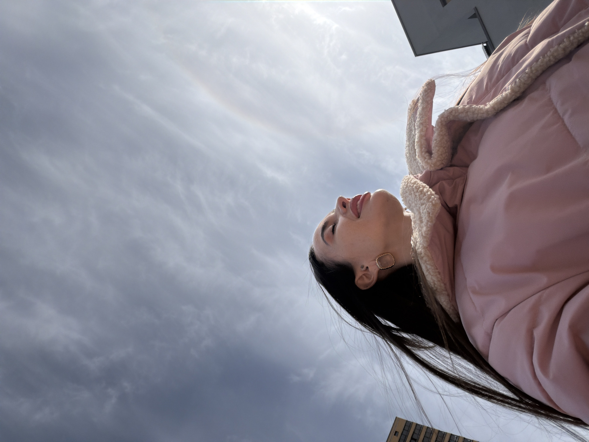 A low-angle shot reveals a woman with long, dark hair and a pink winter jacket, her head tilted back, tongue slightly extended, against a cloudy sky. A building is faintly visible in the corner of the frame. The location is Saint Petersburg, Russia.