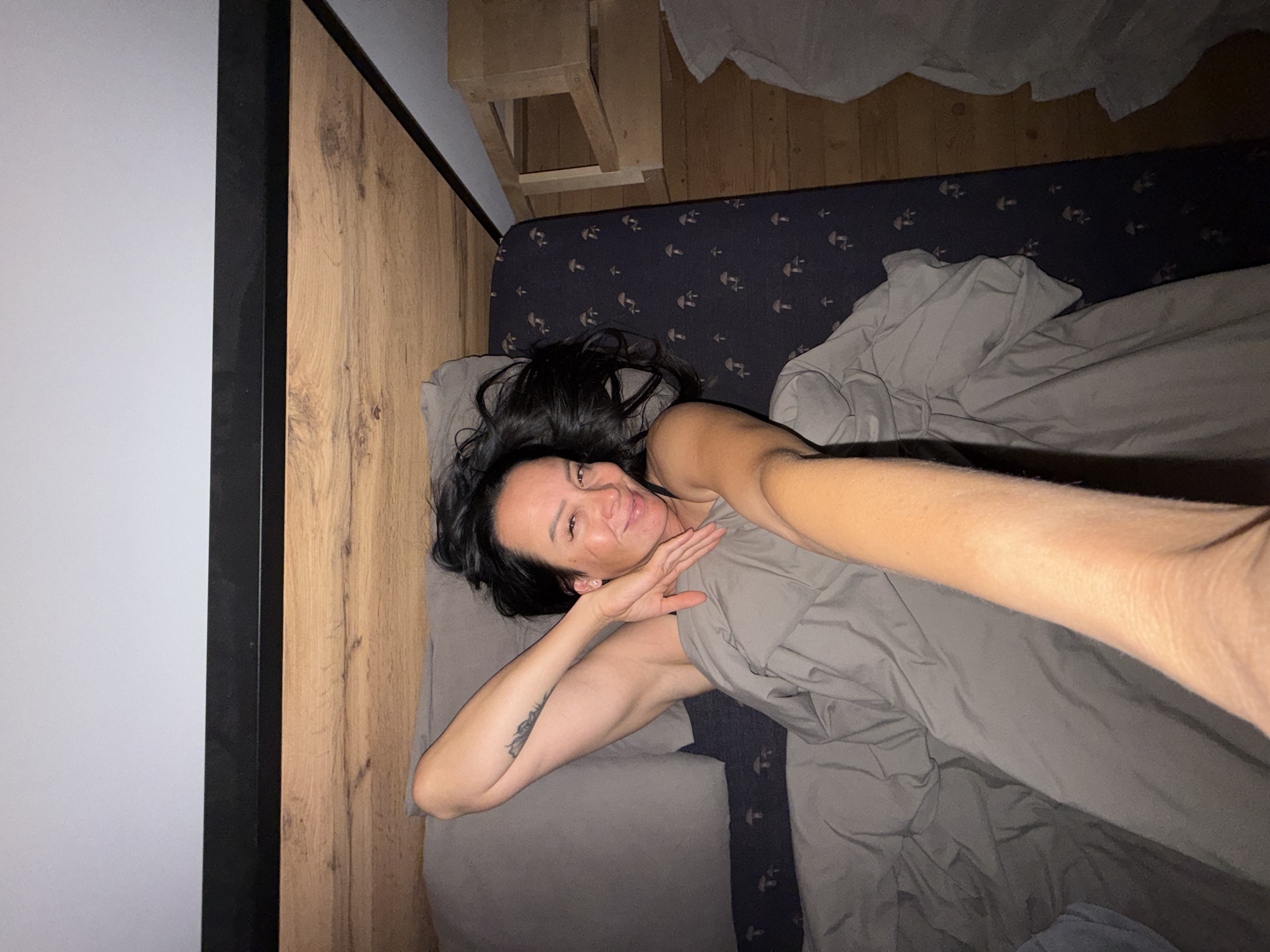 A person with long, dark hair lies in a bed, holding a blanket around them. They are looking towards the camera with a smile and have their left arm raised behind their head. The bed has grey sheets and a dark duvet with a mushroom pattern. The headboard is made of wood, and there is a wooden structure to the right of the bed. The scene is indoors, likely a bedroom.