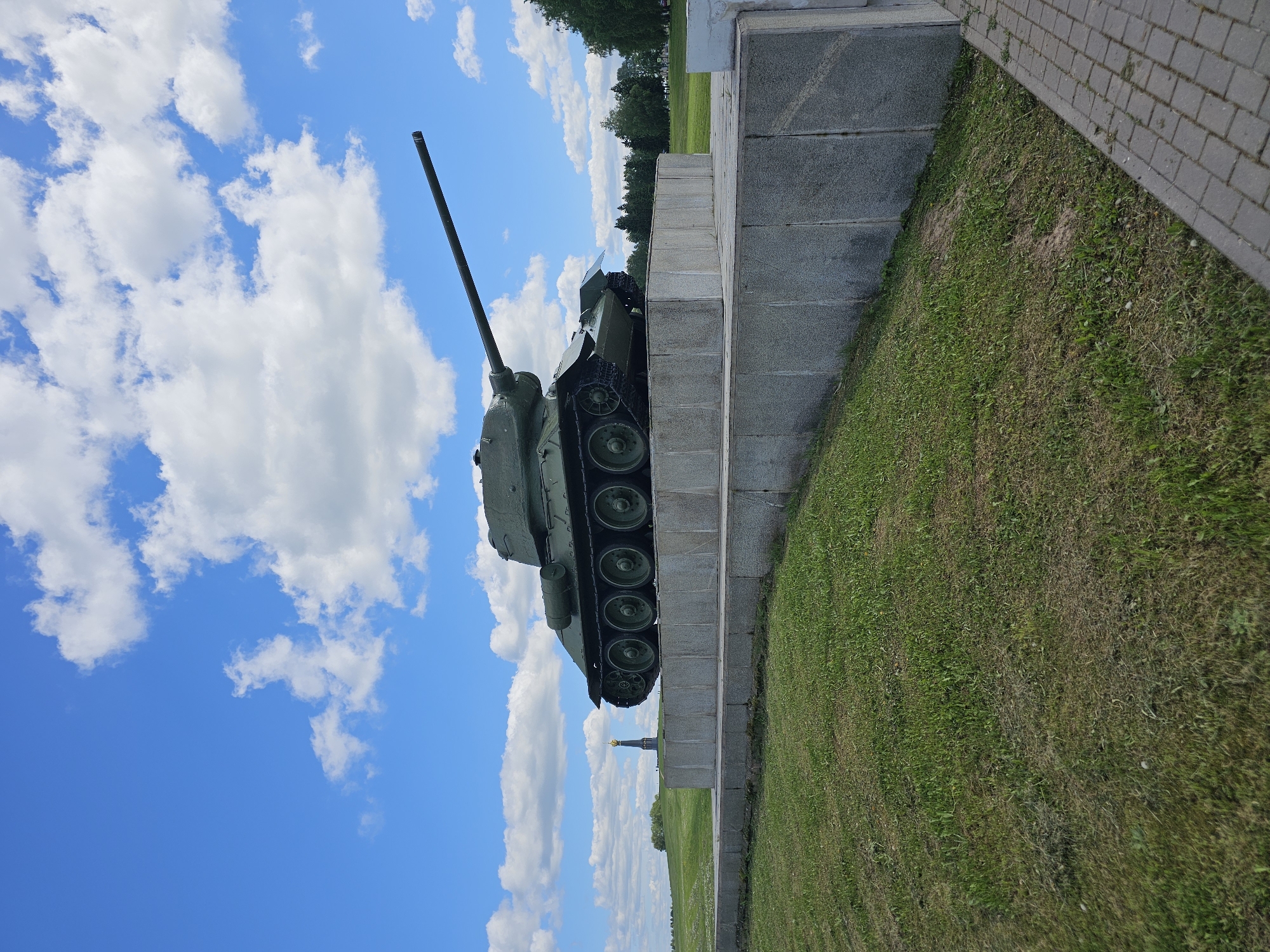 A T-34 tank rests atop a gray stone monument alongside a grassy field under a blue sky scattered with white clouds. A paved walkway borders the grass. The scene is located in Borodino, Russia.