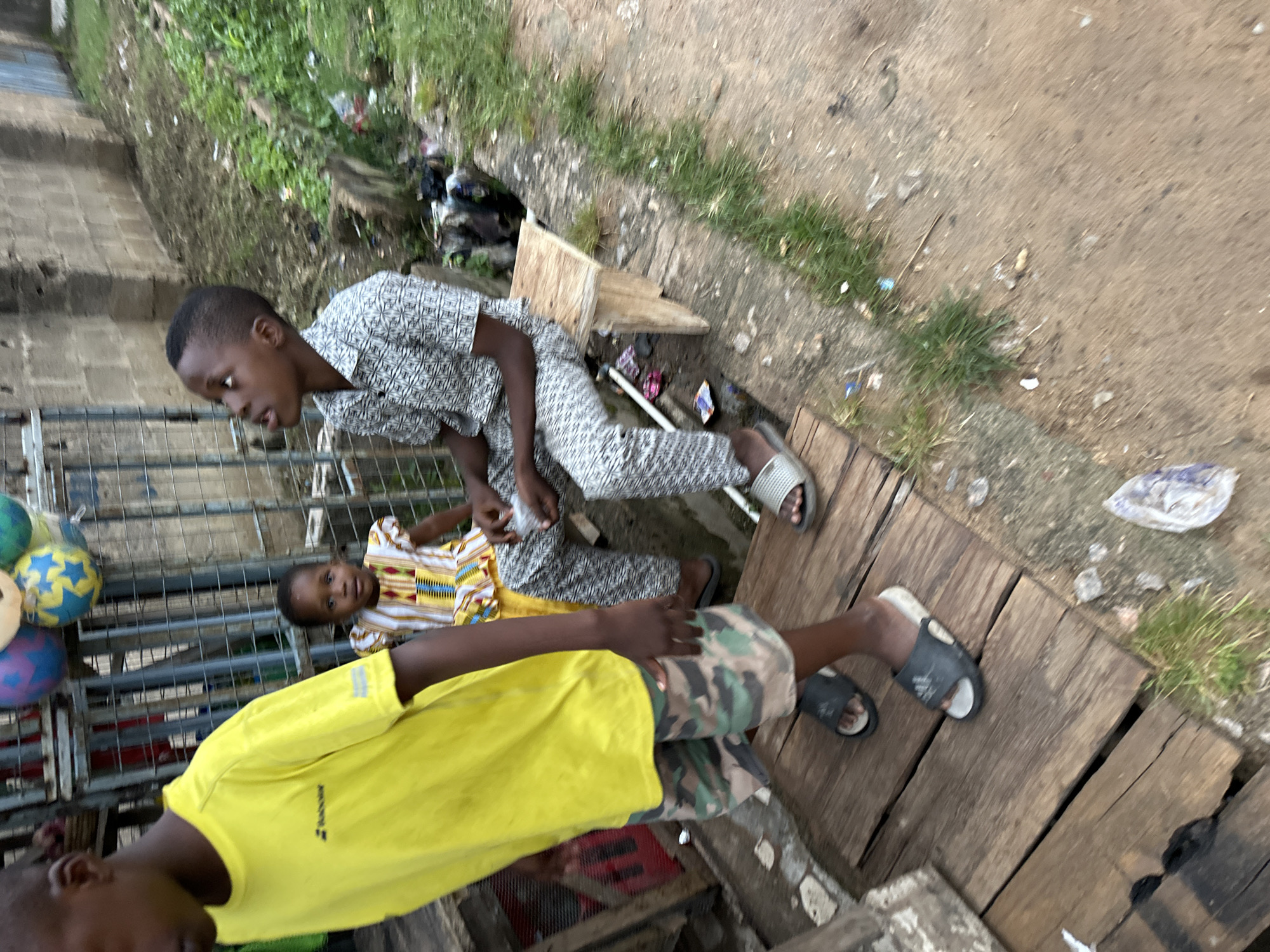 In a setting that appears to be Narayi, Nigeria, a group of four children are present. In the foreground, a young boy wearing a bright yellow t-shirt and camouflage shorts is partially visible, facing away from the camera and walking on a wooden platform. To his right, a young girl in a colorful, patterned dress is seated, looking towards the viewer. Beside her, a boy in a patterned button-down shirt and matching pants is seated on a wooden bench, looking upwards. Behind the seated children, another individual is partially visible, also appearing to be a child. The environment is outdoors, with a brick wall in the background and overgrown vegetation. Debris is scattered on the ground. The overall impression is a street scene with children in a somewhat cluttered urban setting.