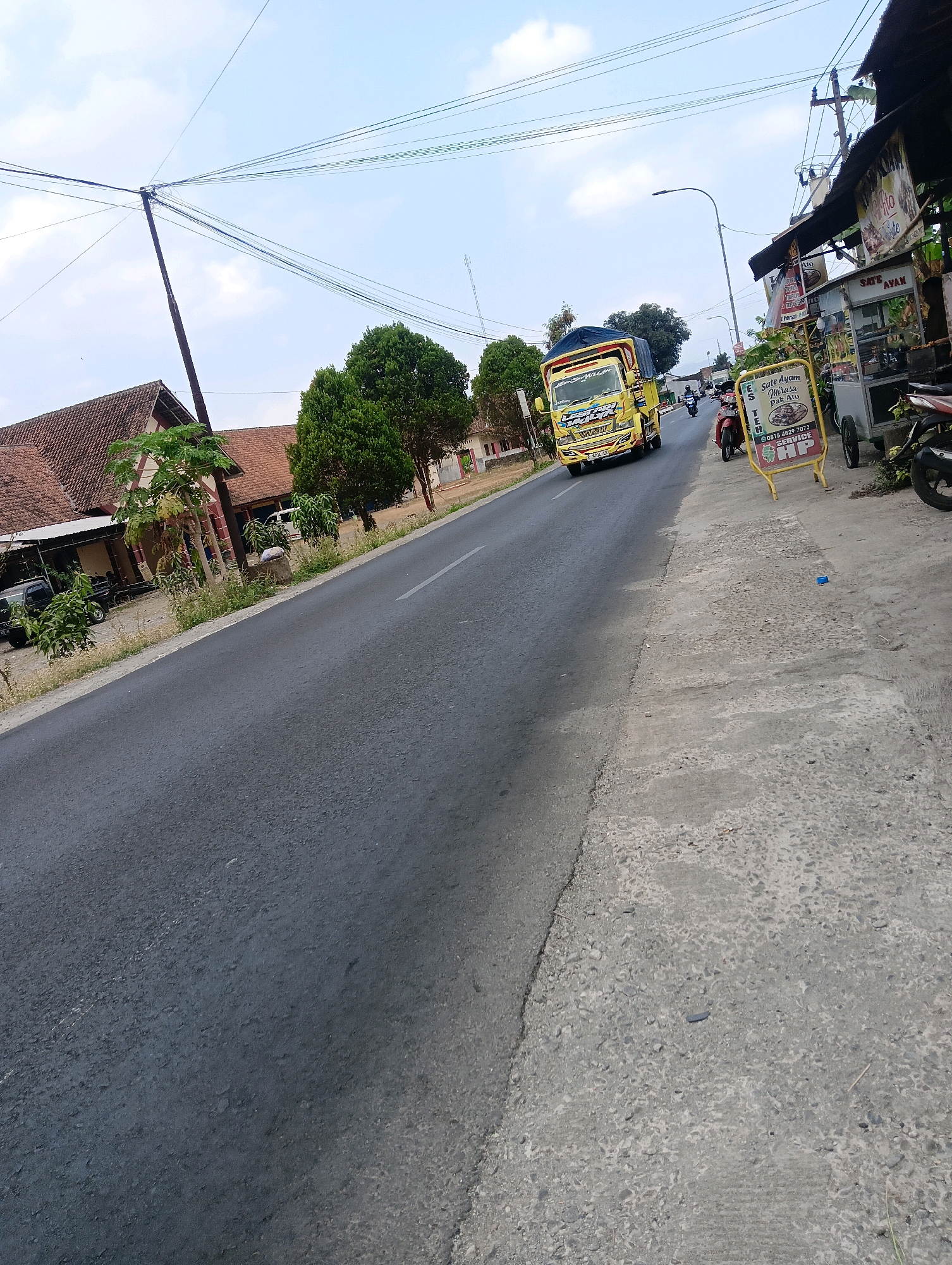 The image depicts a street scene in Sokaraja, Indonesia. On the right side of the road, there is a food stall with a yellow sign advertising "SATE AYAM" (Chicken Satay) and a phone number for service. A motorcycle is parked to the right of the stall. Further down the road, a yellow truck is in motion. On the left side of the road, there are houses with tiled roofs and some trees. Overhead, a network of power lines is visible against a partly cloudy sky. The road surface appears to be asphalt with some patches of concrete on the side.